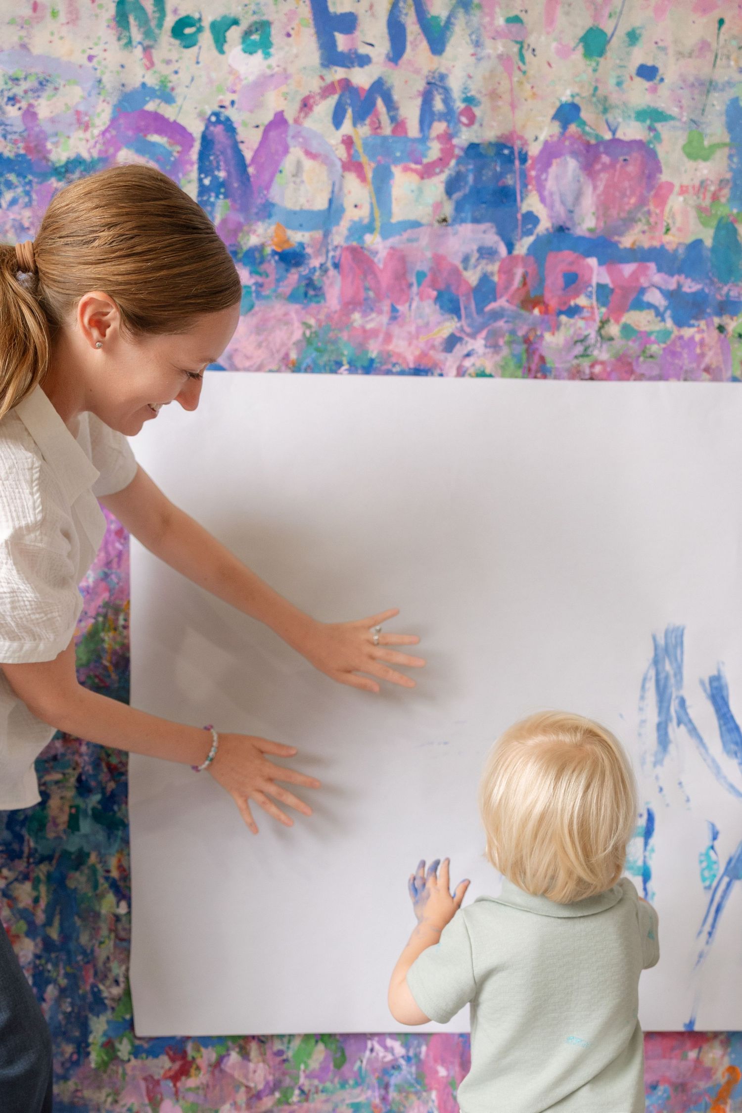 Woman and toddler doing finger painting on large white canvas against colorful painted wall.
