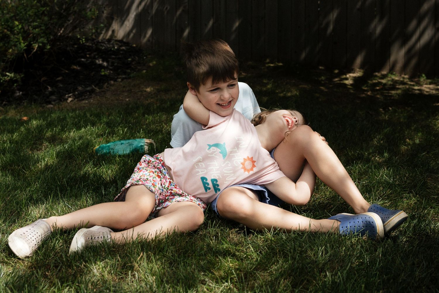 Two children sitting together on green grass in a sunny backyard, smiling and hugging outdoors.