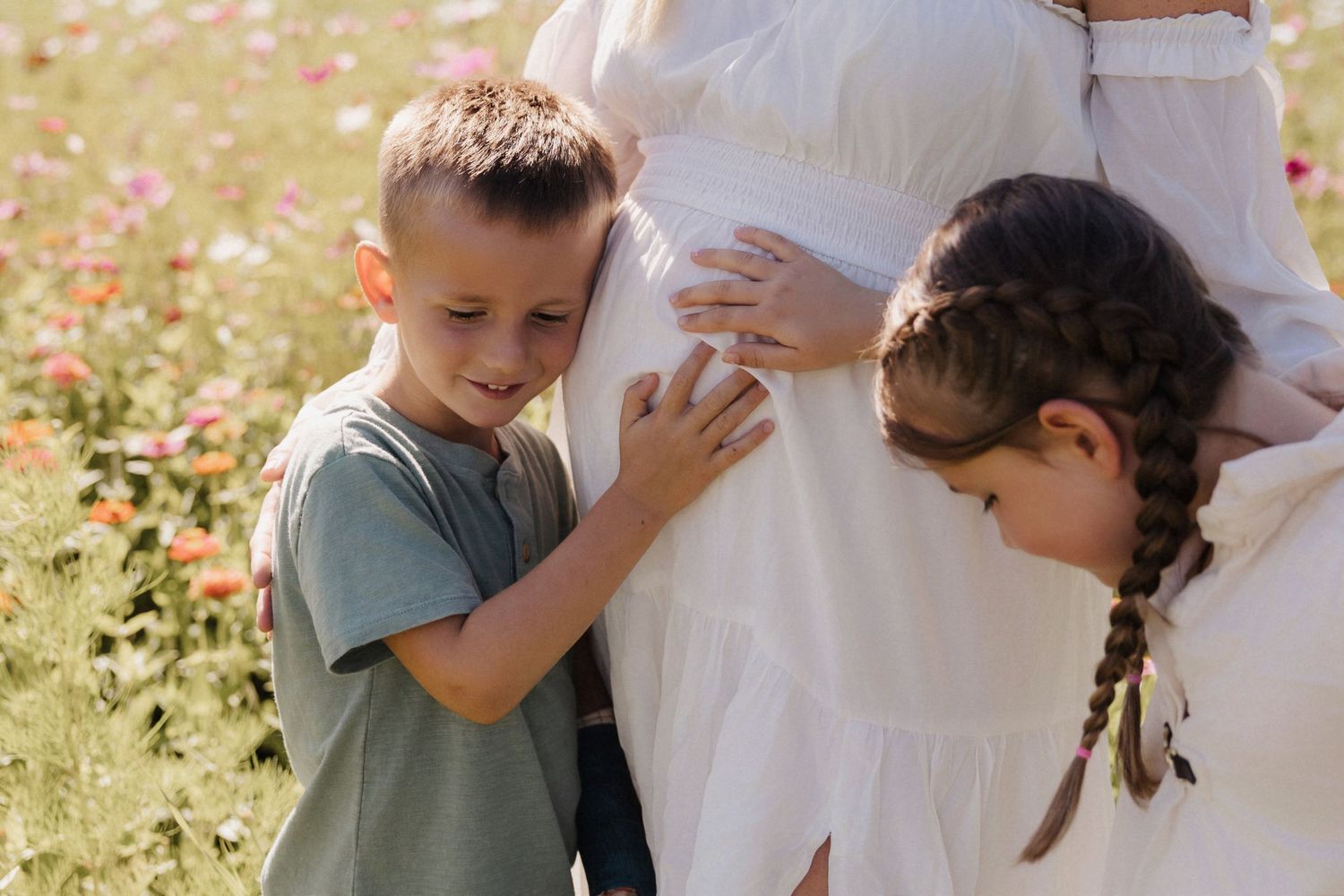 Two children touch a pregnant woman's belly in a flower field.