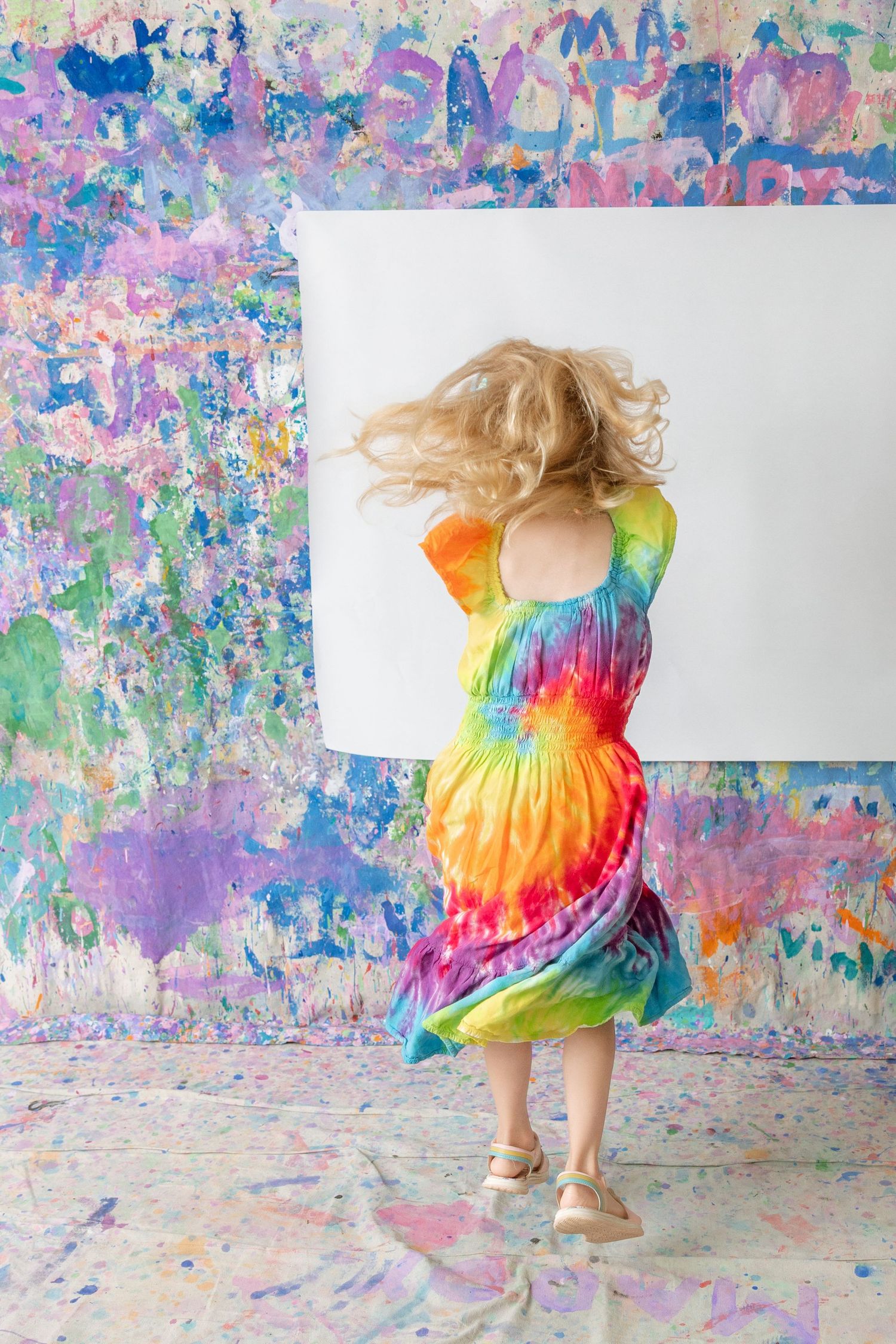 A young girl in a colorful tie-dye dress dances in front of a paint-splattered wall and canvas.