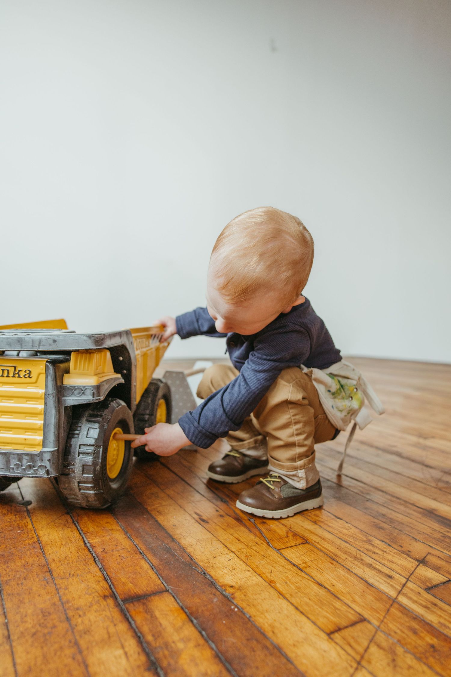 Blonde toddler boy playing with yellow Tonka truck on hardwood floor.