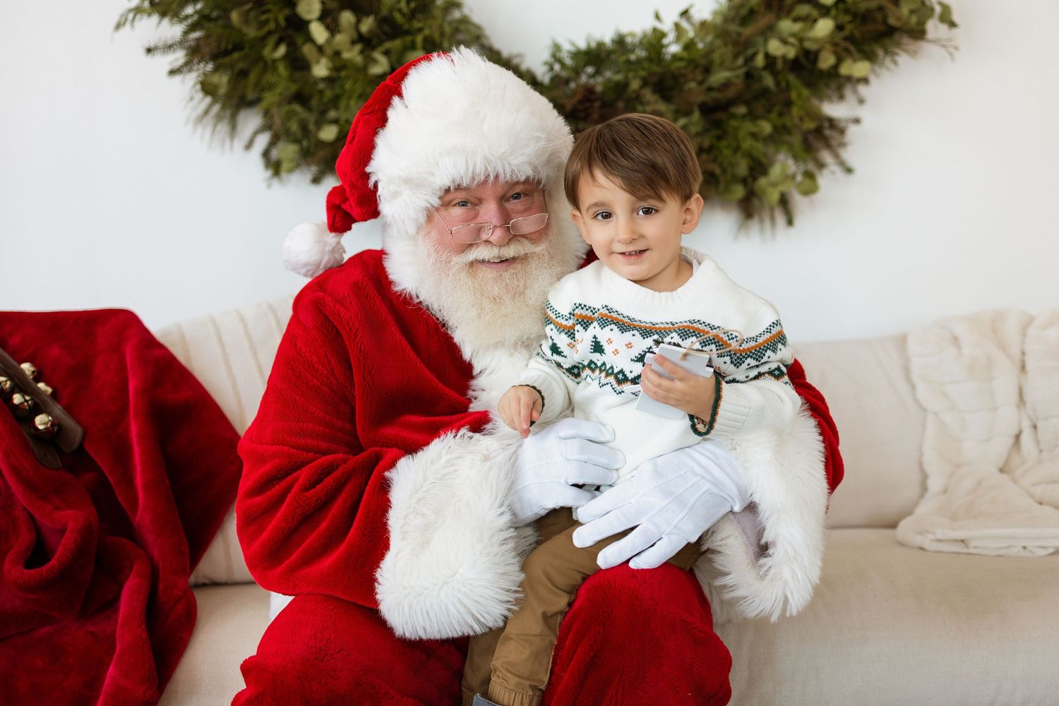 Santa Claus sits on a cream sofa holding a young boy in a Nordic sweater near a Christmas wreath.