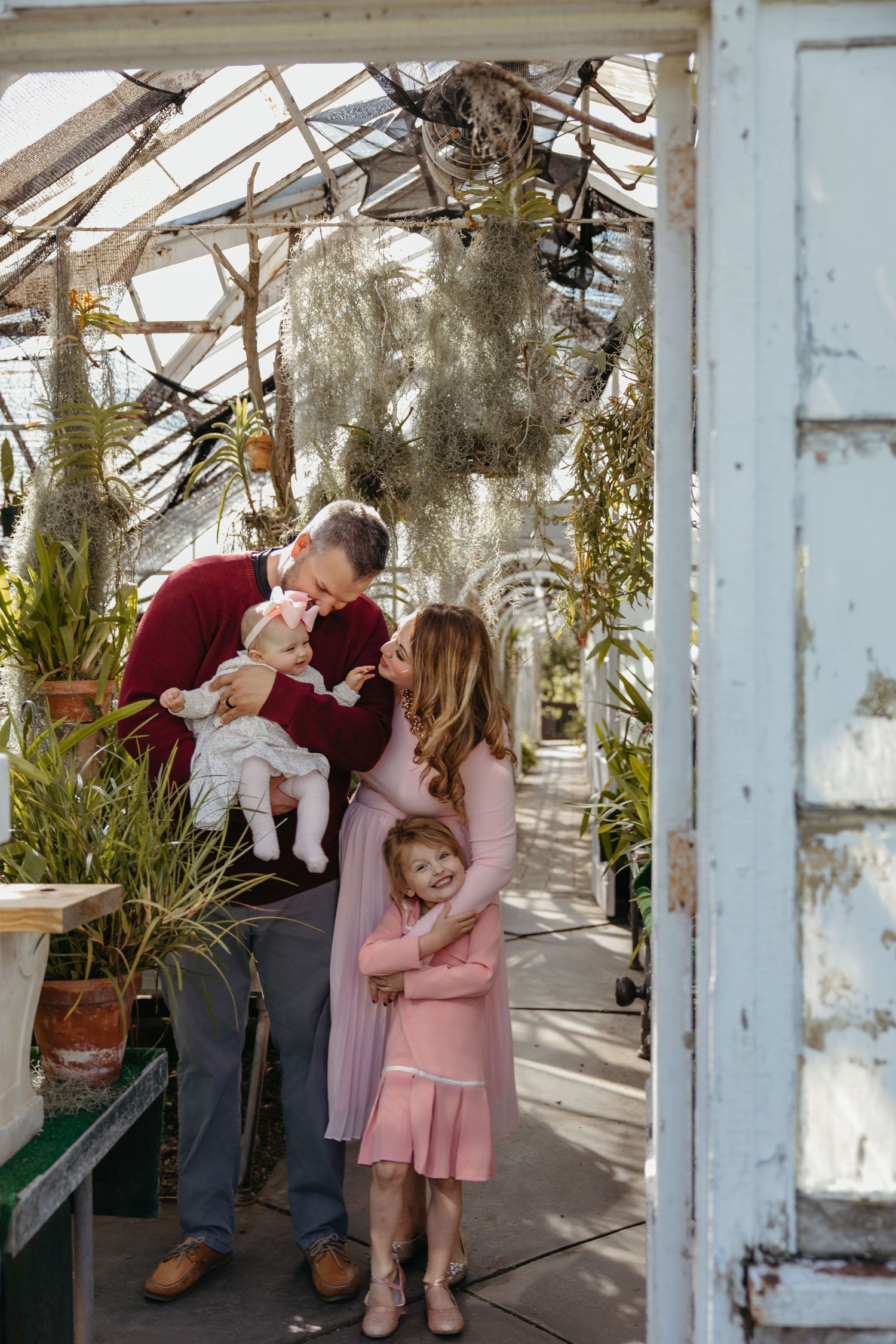 Family sharing a tender moment in a lush greenhouse with two young daughters in pink dresses.