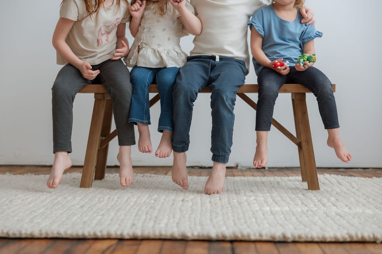 Four barefoot children sitting on wooden benches with feet dangling over a white rug.