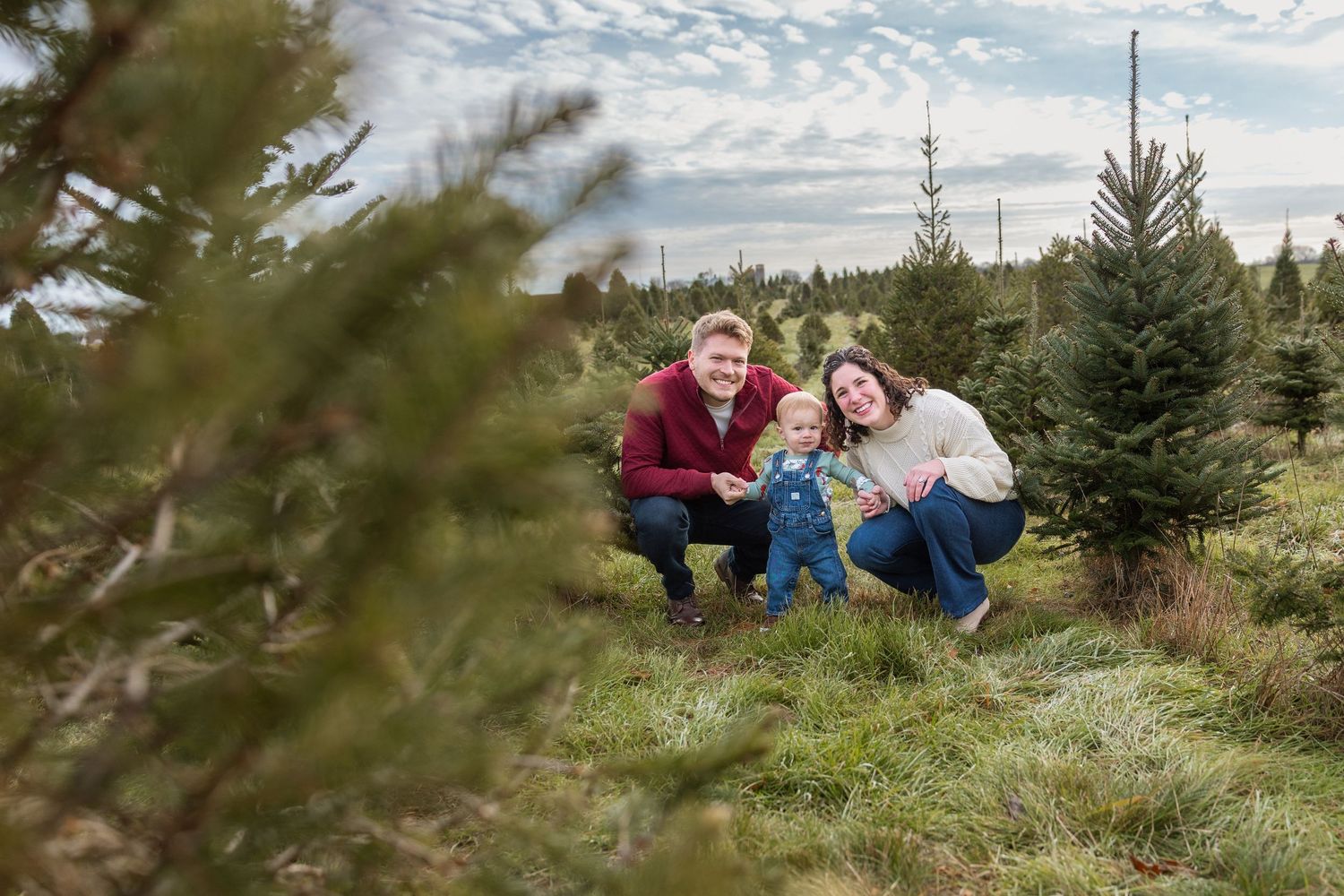 Family of three posing among Christmas trees at a tree farm, parents crouching beside toddler on a cloudy winter day.