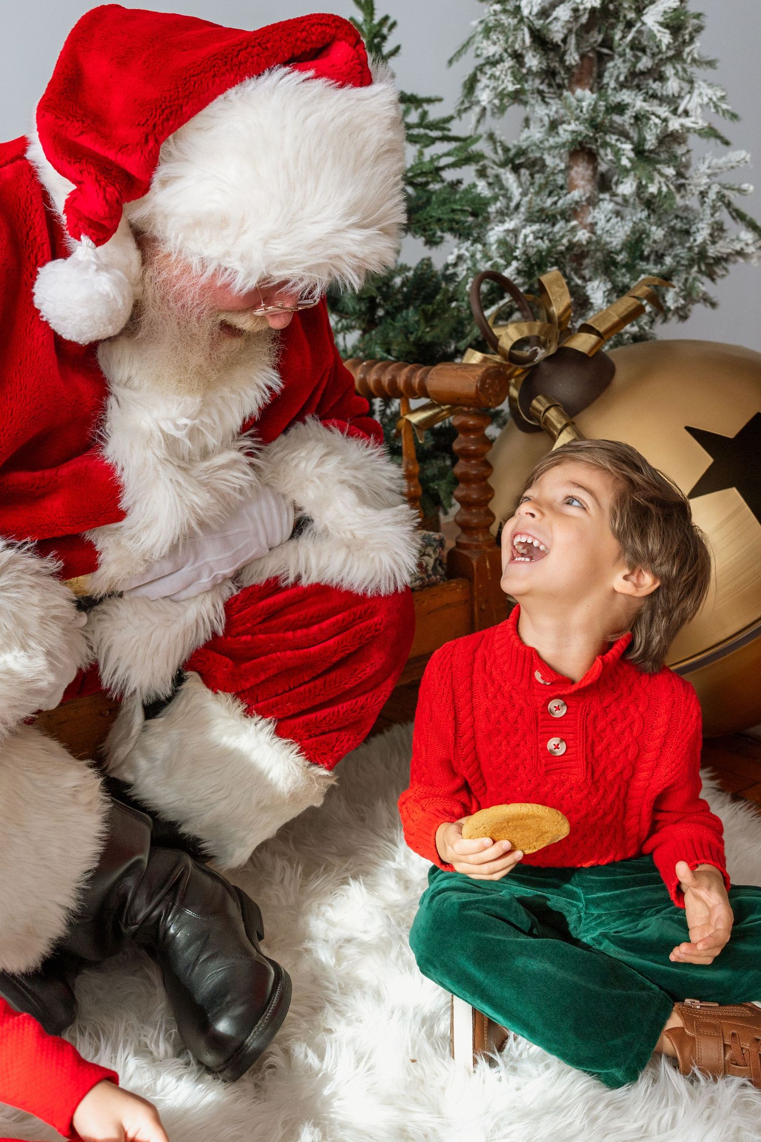 A joyful boy in a red sweater laughing with Santa Claus during a Christmas portrait session.