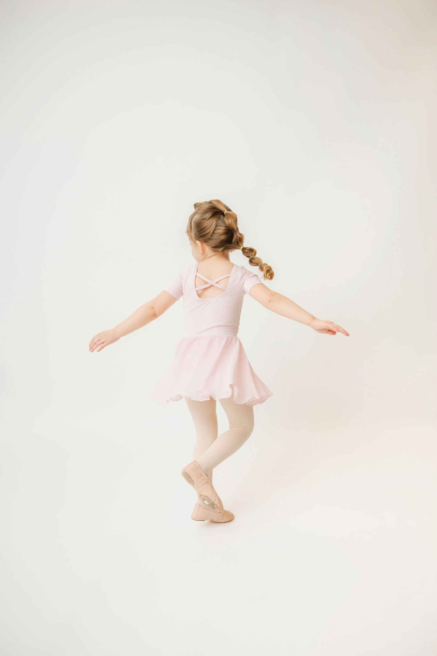 Young girl in pink ballet leotard and skirt dancing gracefully with arms outstretched, viewed from behind.