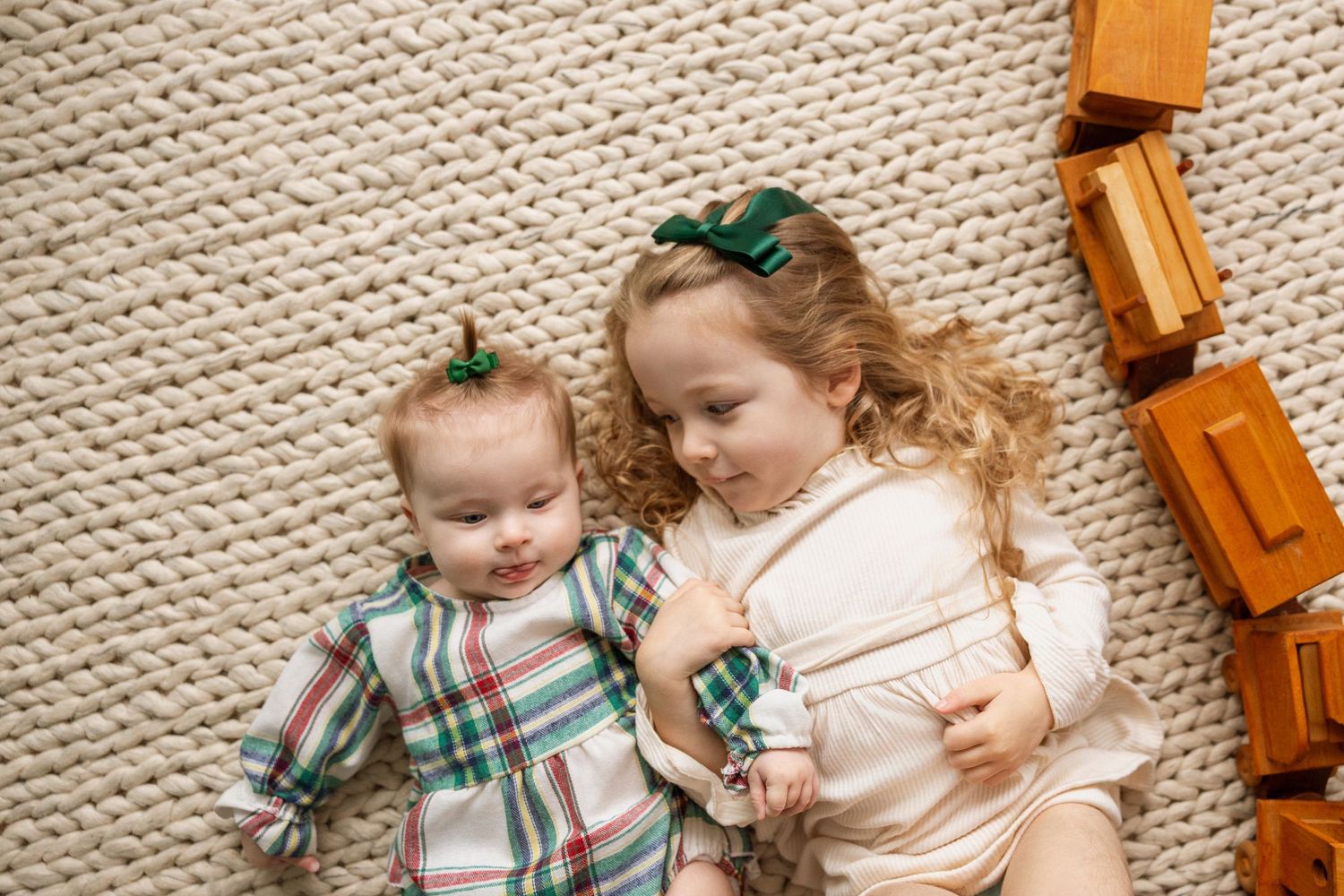 Two young siblings lie on a knit rug, the older curly-haired girl hugging her baby sibling surrounded by wooden blocks.