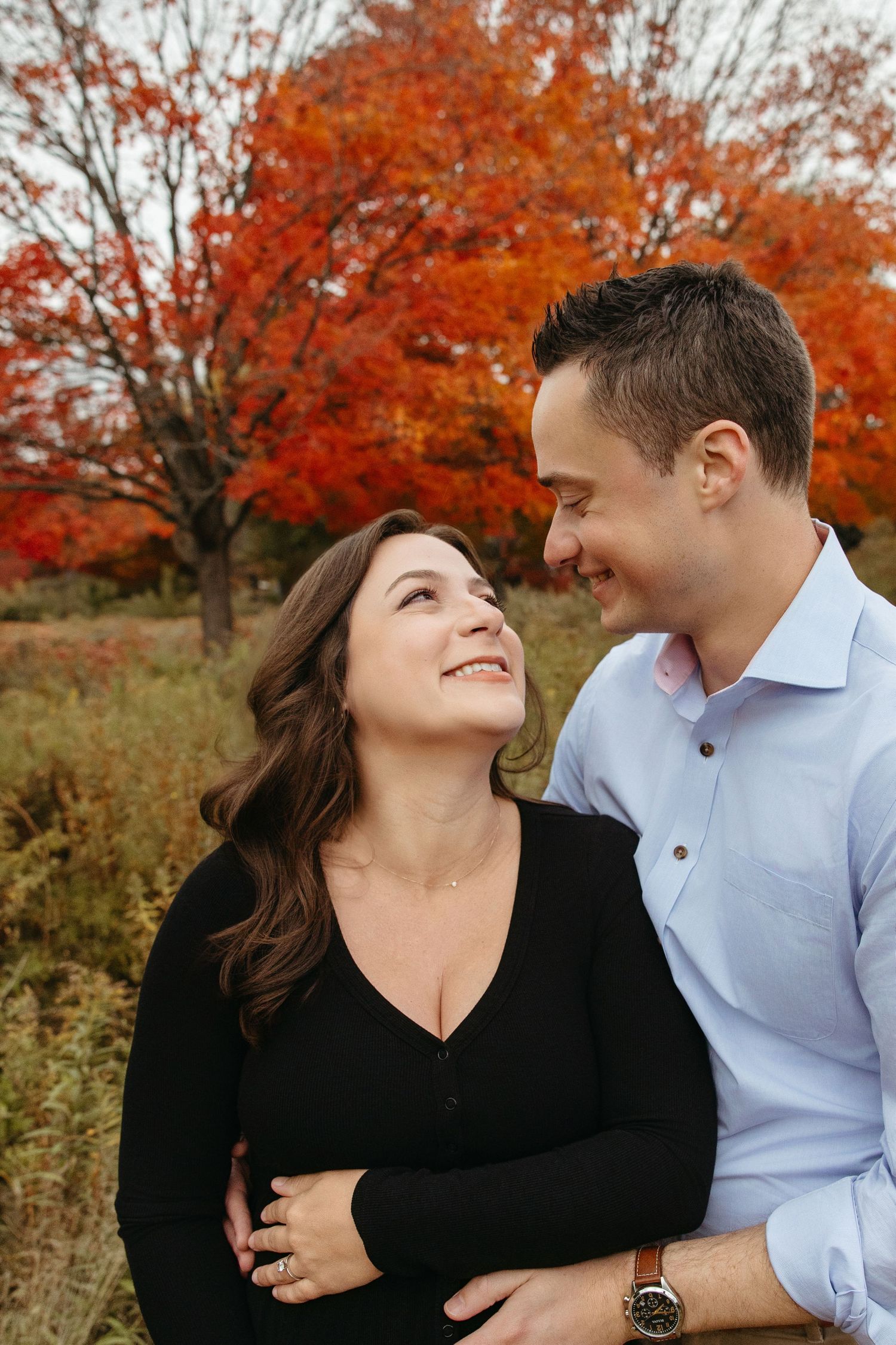 Smiling couple embracing in front of a vibrant red maple tree during autumn season.