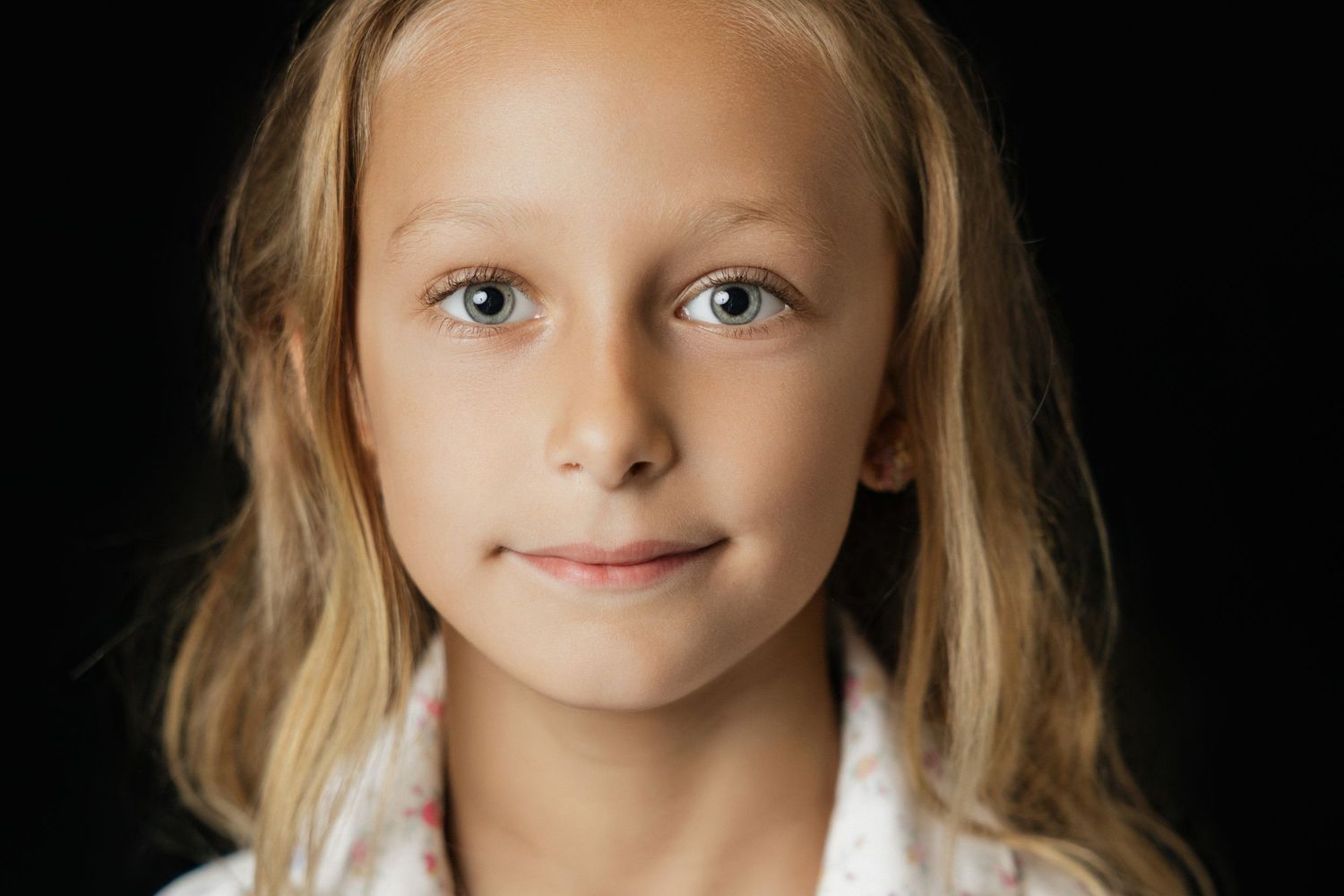 Portrait of a smiling young blonde girl with blue eyes against a dark background.