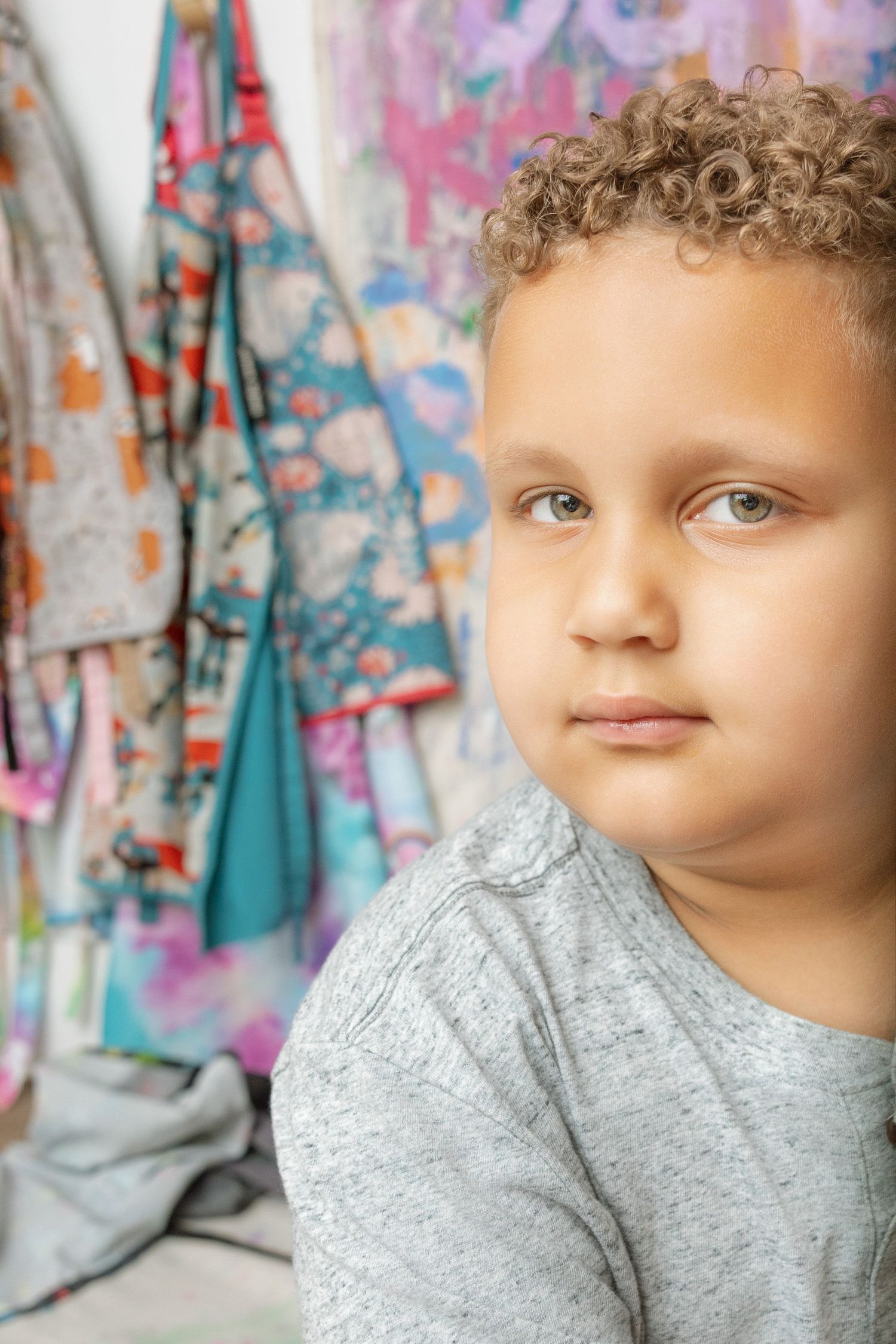A young boy with curly blonde hair wearing a gray shirt sits in front of colorful hanging fabric.