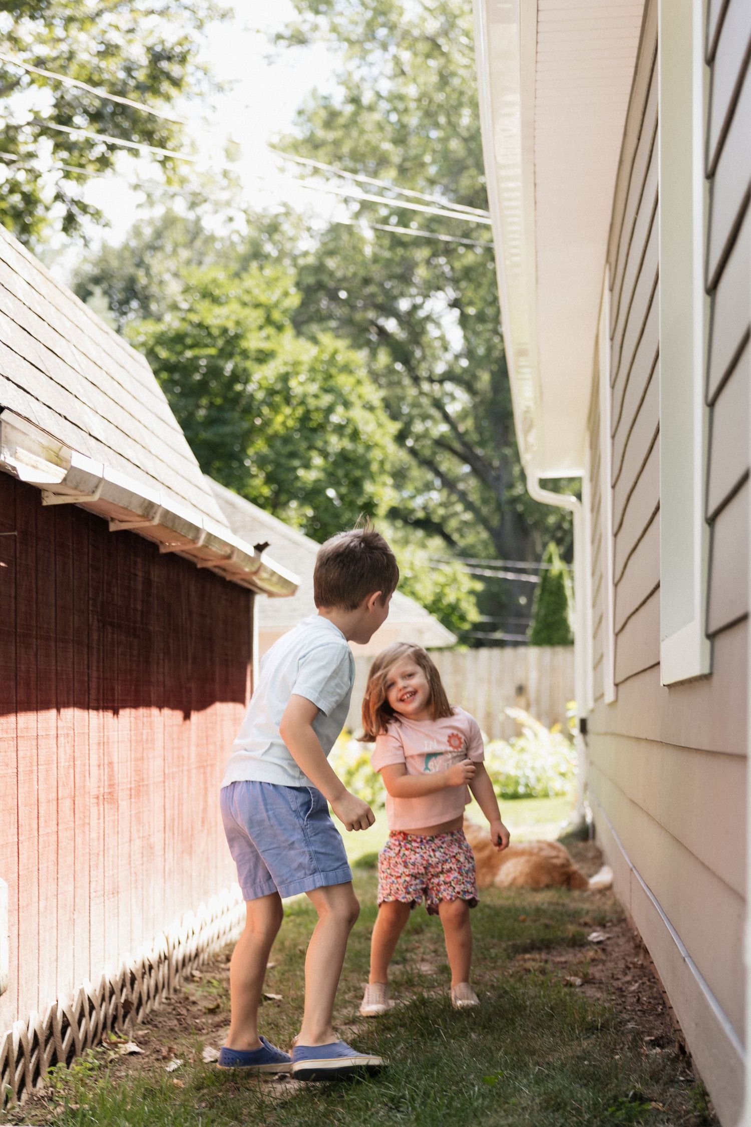 Two young children playing and laughing together in a sunny backyard between two houses.