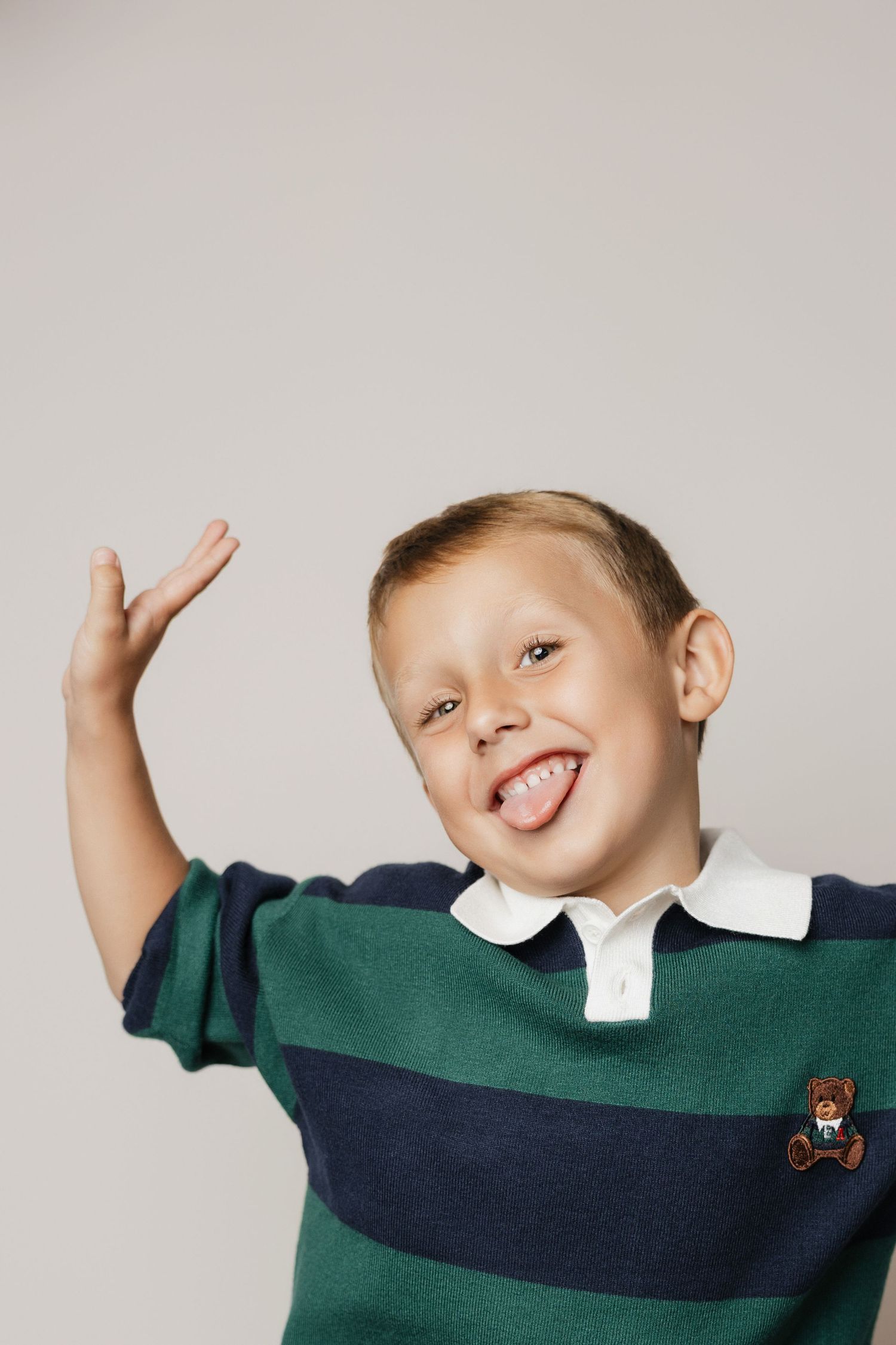 Young boy in striped green polo shirt sticking out tongue and raising hand playfully against gray background.