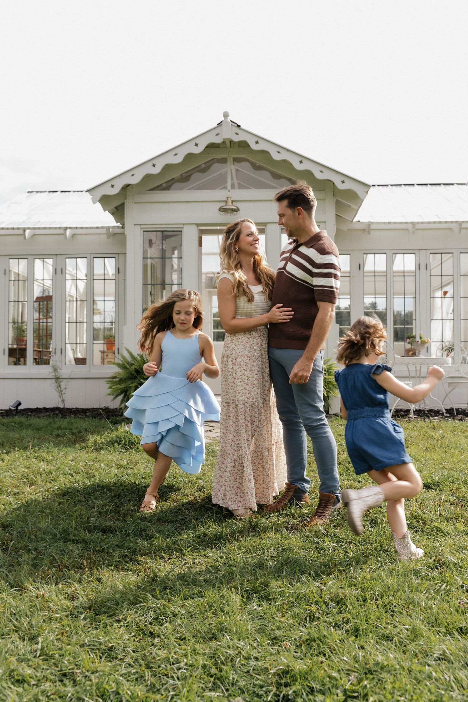 A happy family of four enjoys a sunny day on green grass in front of a charming white greenhouse.