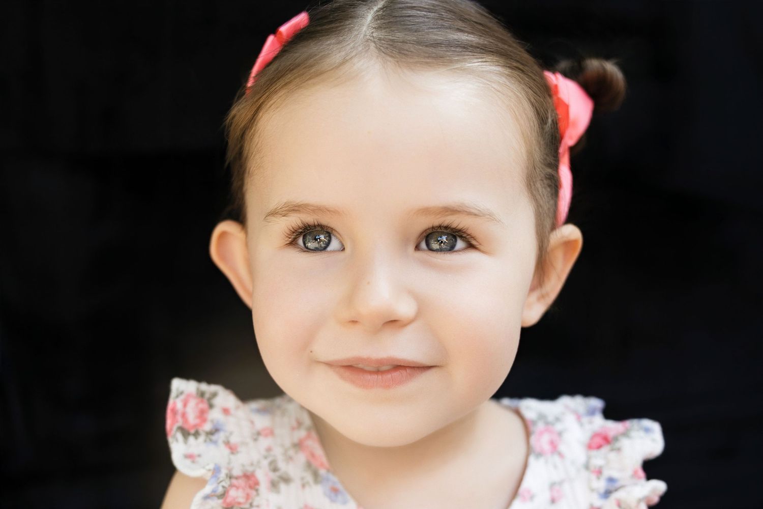 Adorable toddler girl with dark hair in pigtails wearing a floral dress, smiling sweetly at the camera.