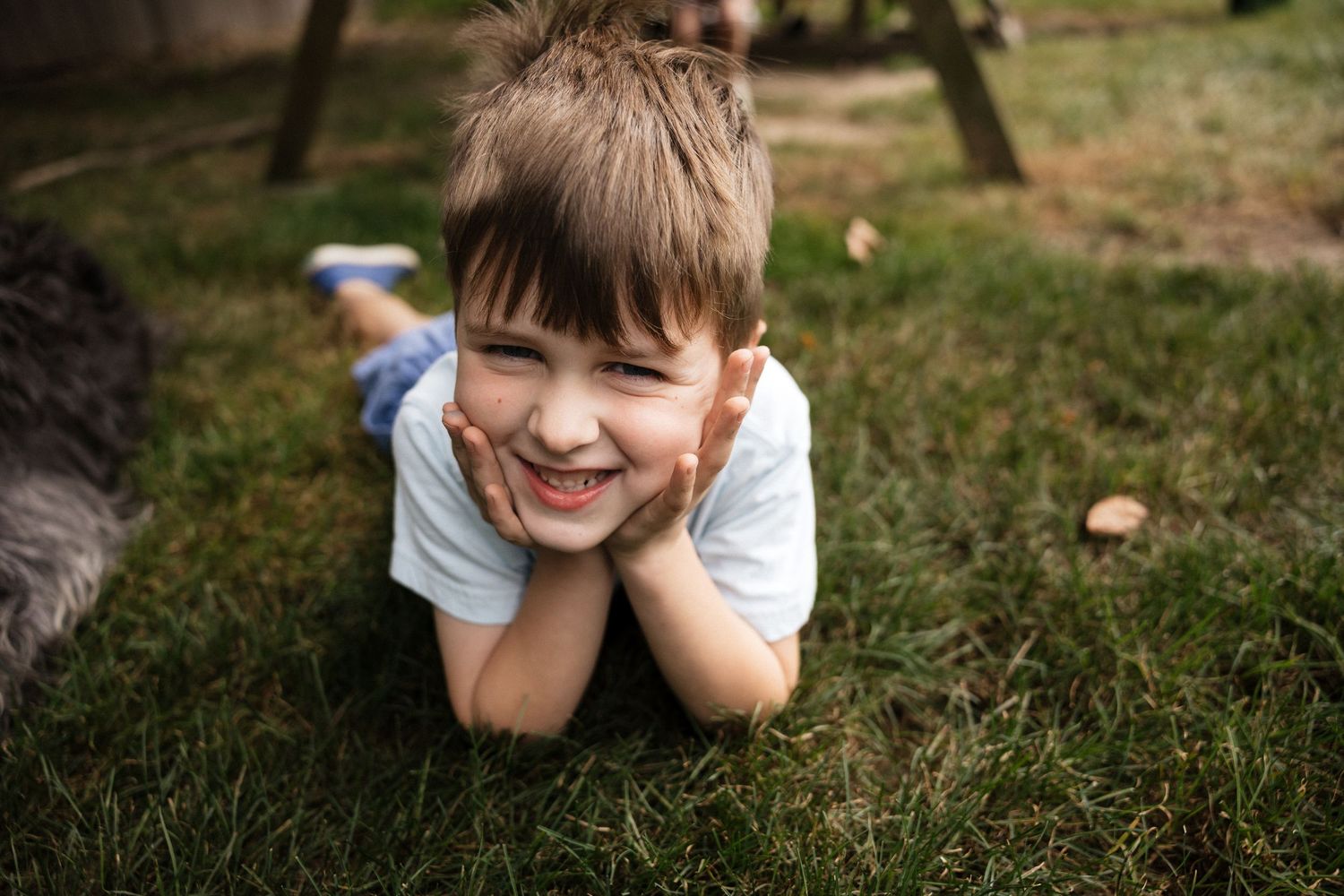 Happy young boy lying on green grass outdoors, smiling with hands on cheeks in a sunny backyard.