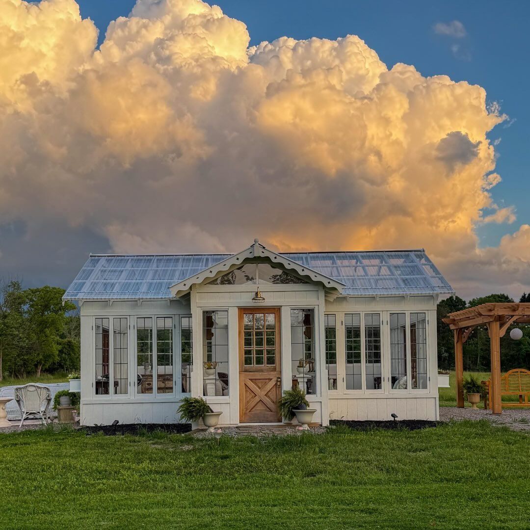 Charming white greenhouse cottage with glass roof panels sits on green lawn beneath dramatic golden sunset clouds.