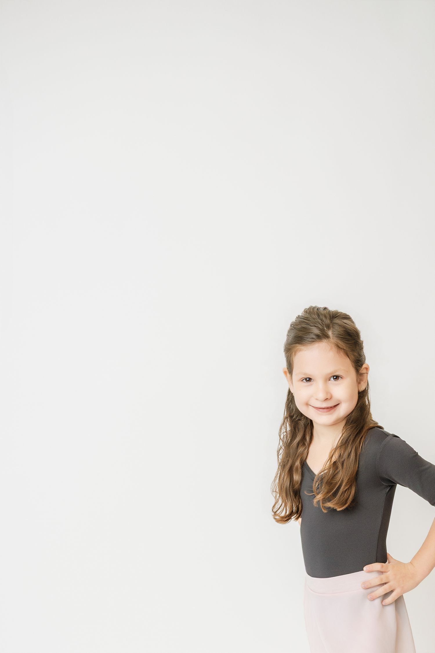 Young girl dancer in gray leotard and pink skirt smiles with hand on hip in a white studio.