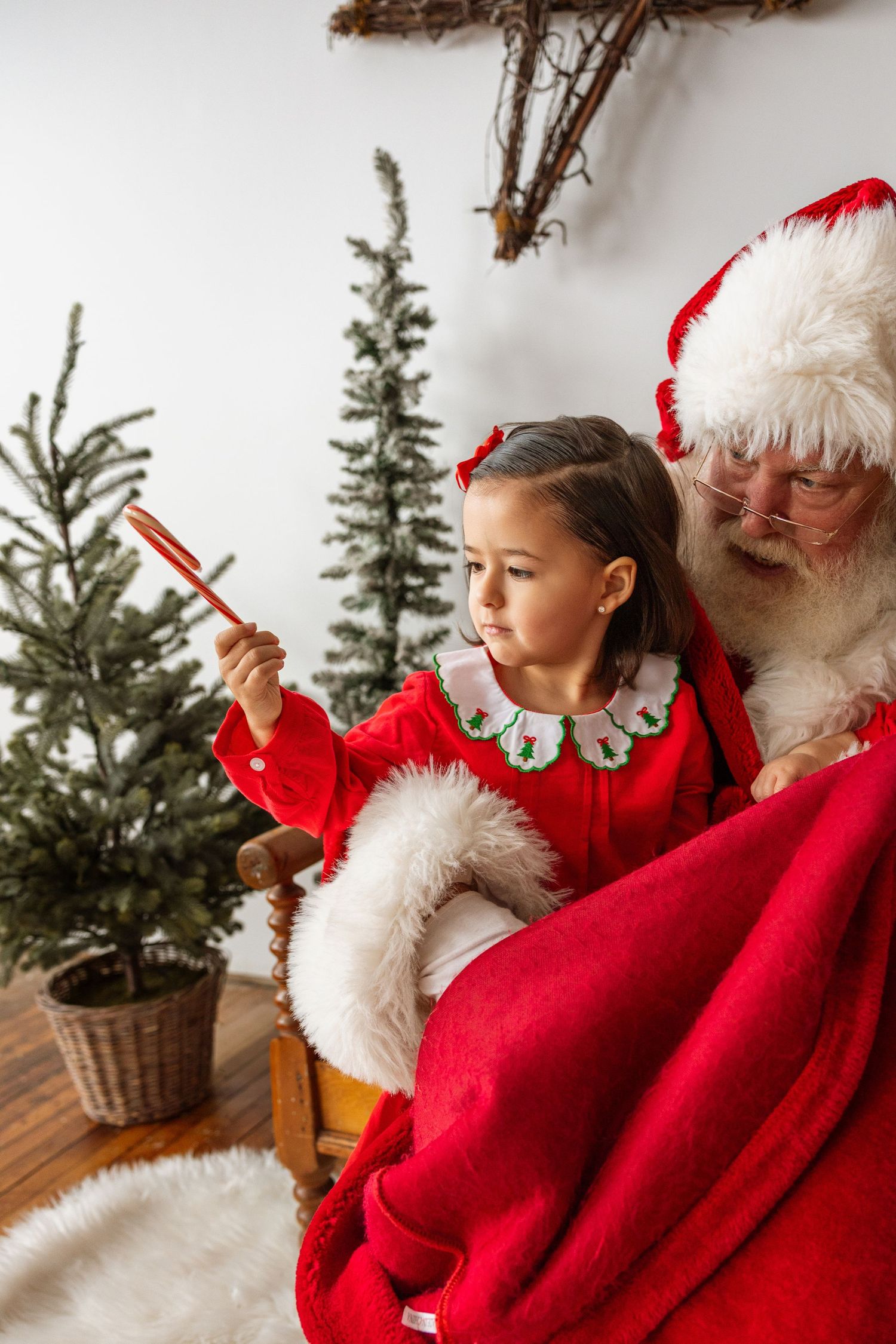 A young girl in a red holiday dress sits on Santa's lap holding a candy cane near Christmas trees.