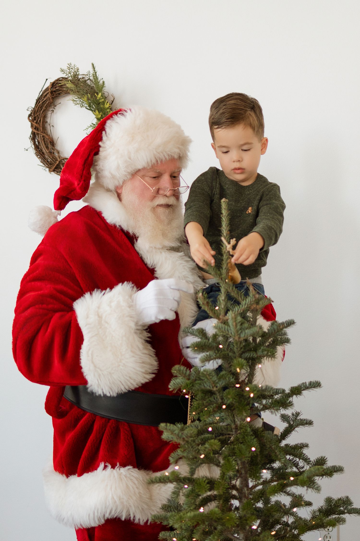 Santa Claus assists a young boy in decorating a small Christmas tree with ornaments indoors.