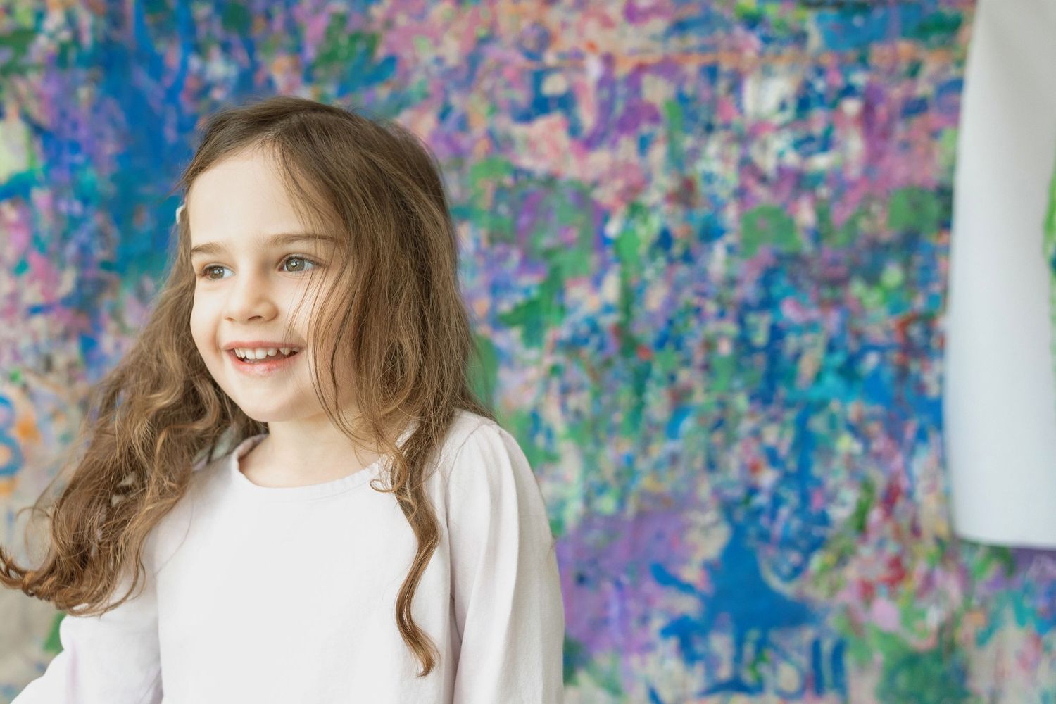 A smiling young girl with long brown hair stands in front of a colorful painted wall.