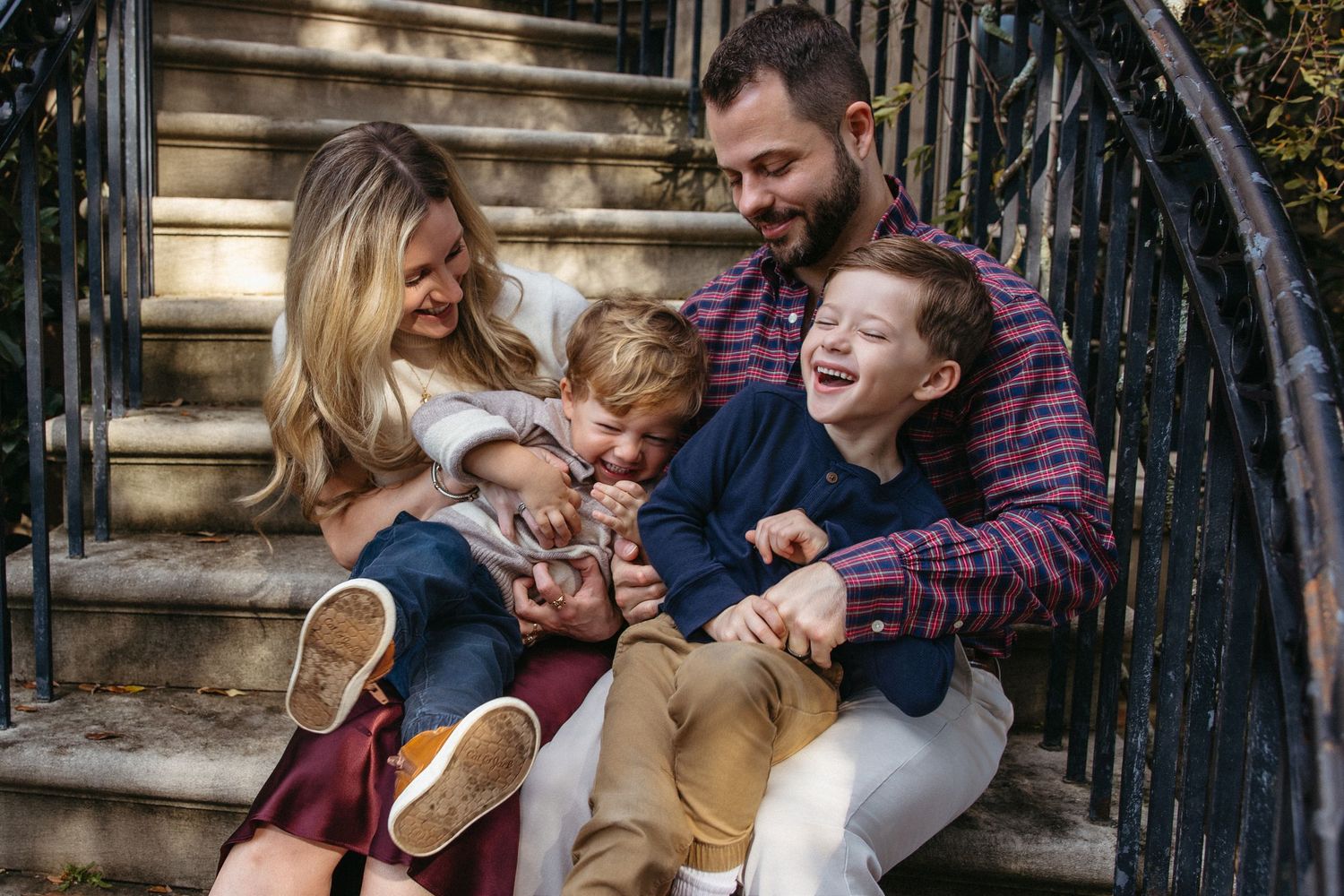 A joyful family of four laughs together on outdoor steps surrounded by greenery.