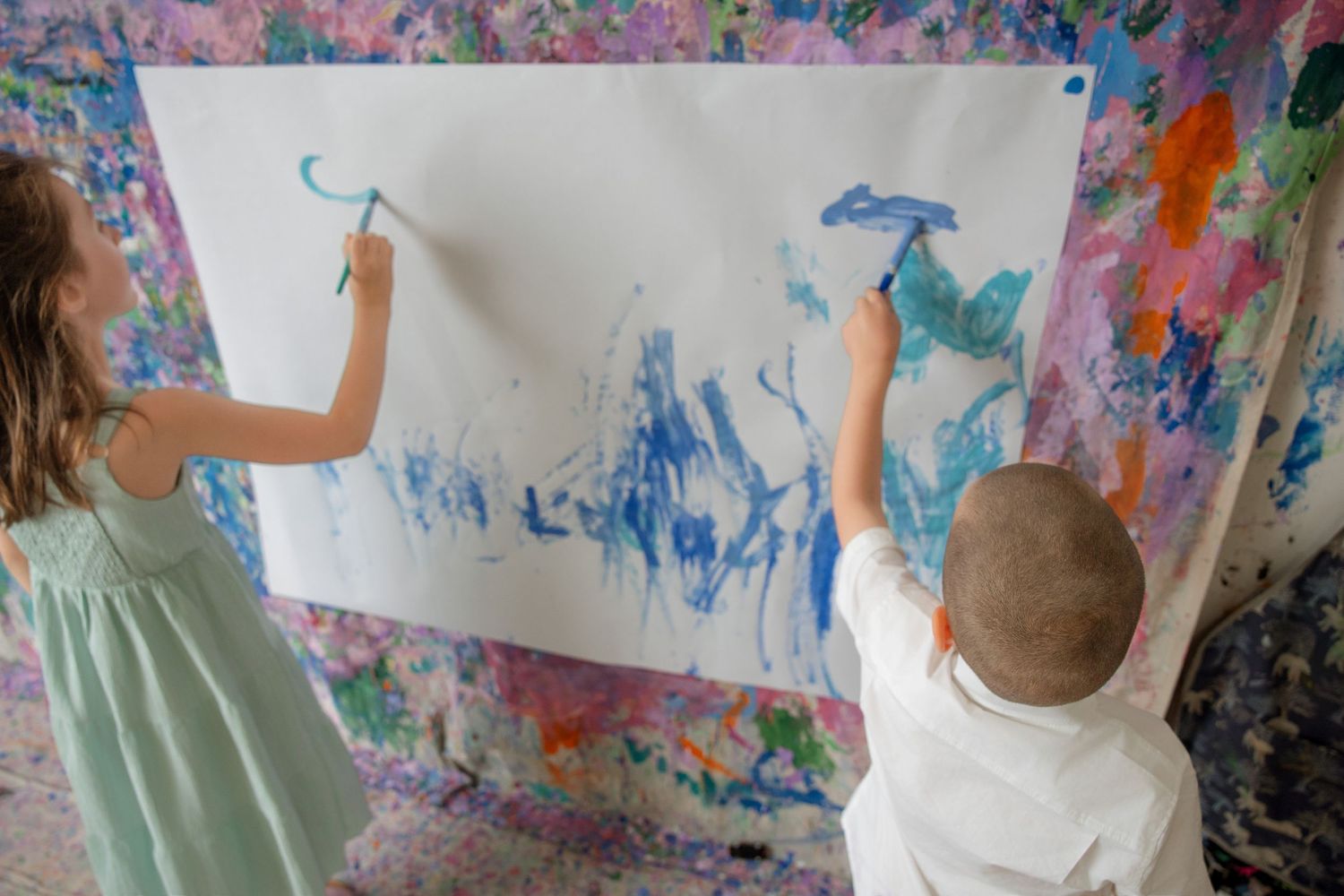 Two children painting on a large white canvas mounted on a colorful splattered wall.
