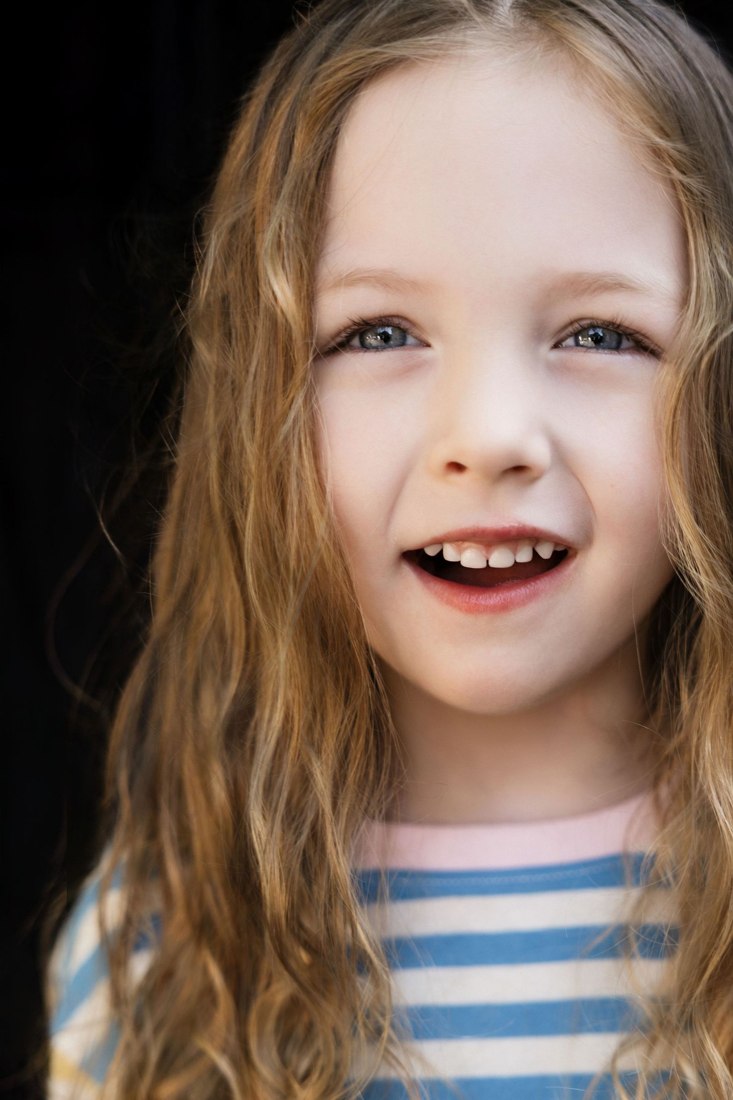 Smiling young girl with long wavy blonde hair and blue eyes wearing a striped shirt against dark background.