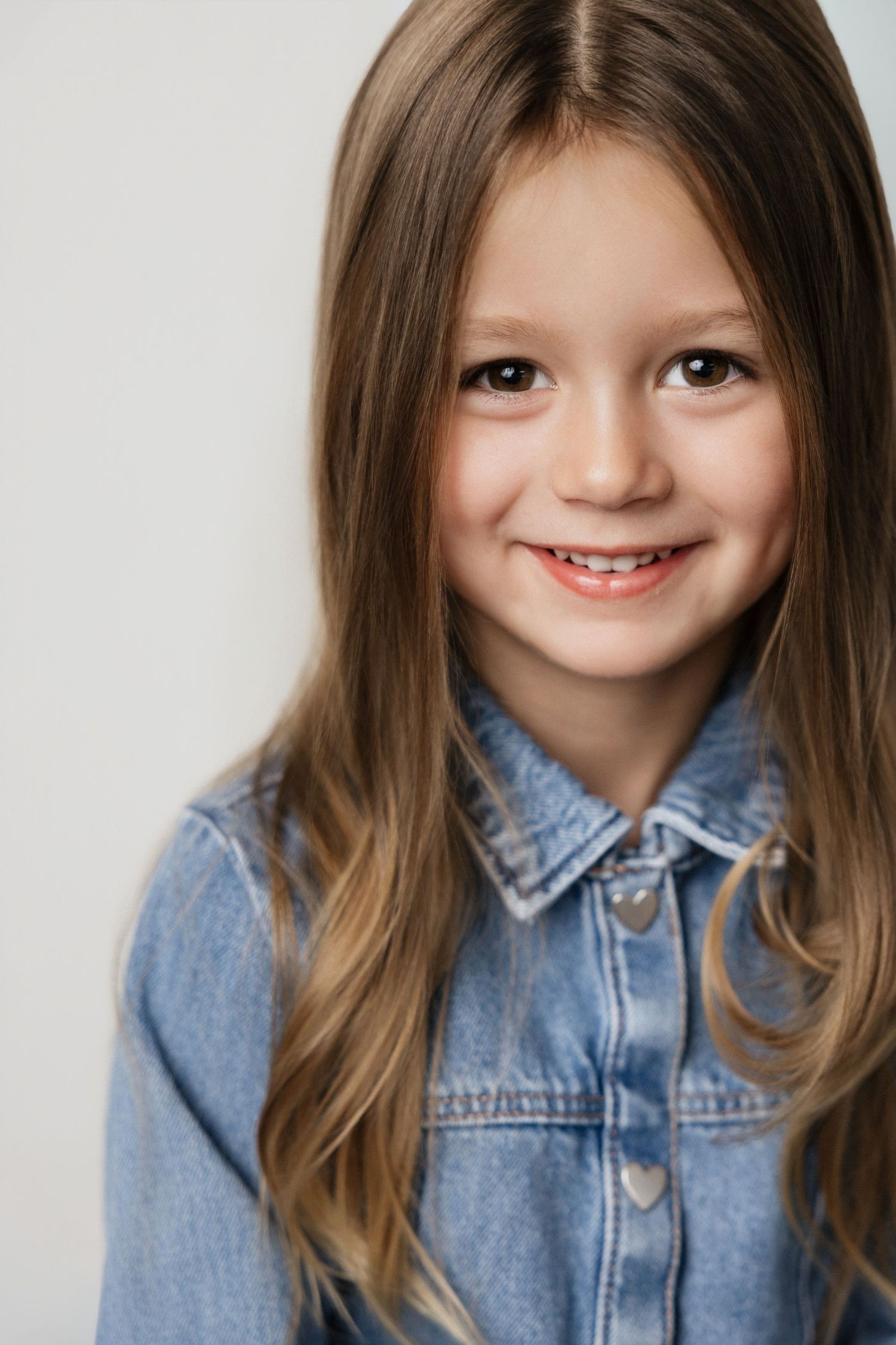 Smiling young girl with long brown hair wearing a denim jacket poses for a portrait photo.