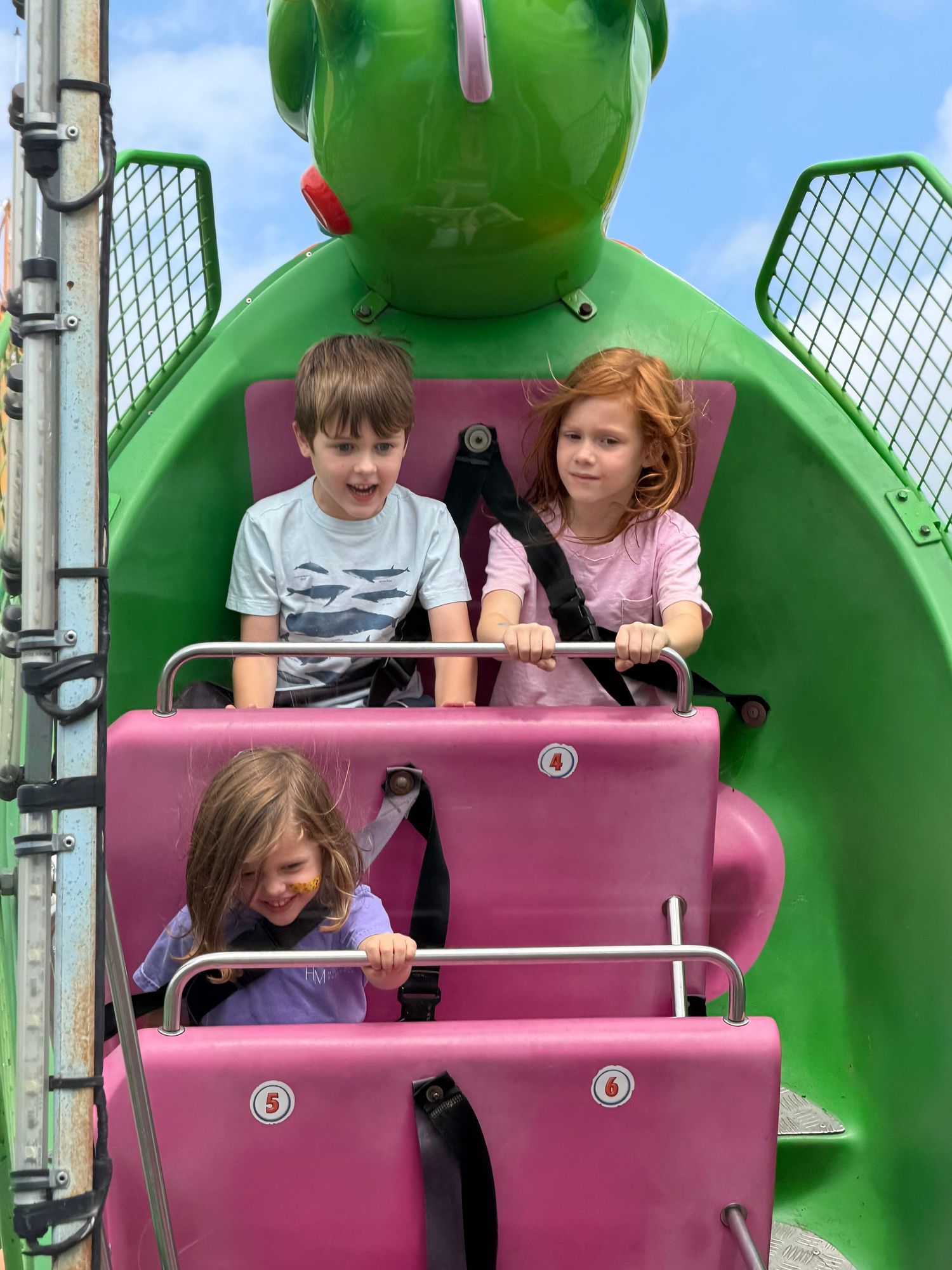 Three children riding a green dragon-themed pink roller coaster ride at an amusement park on a sunny day.