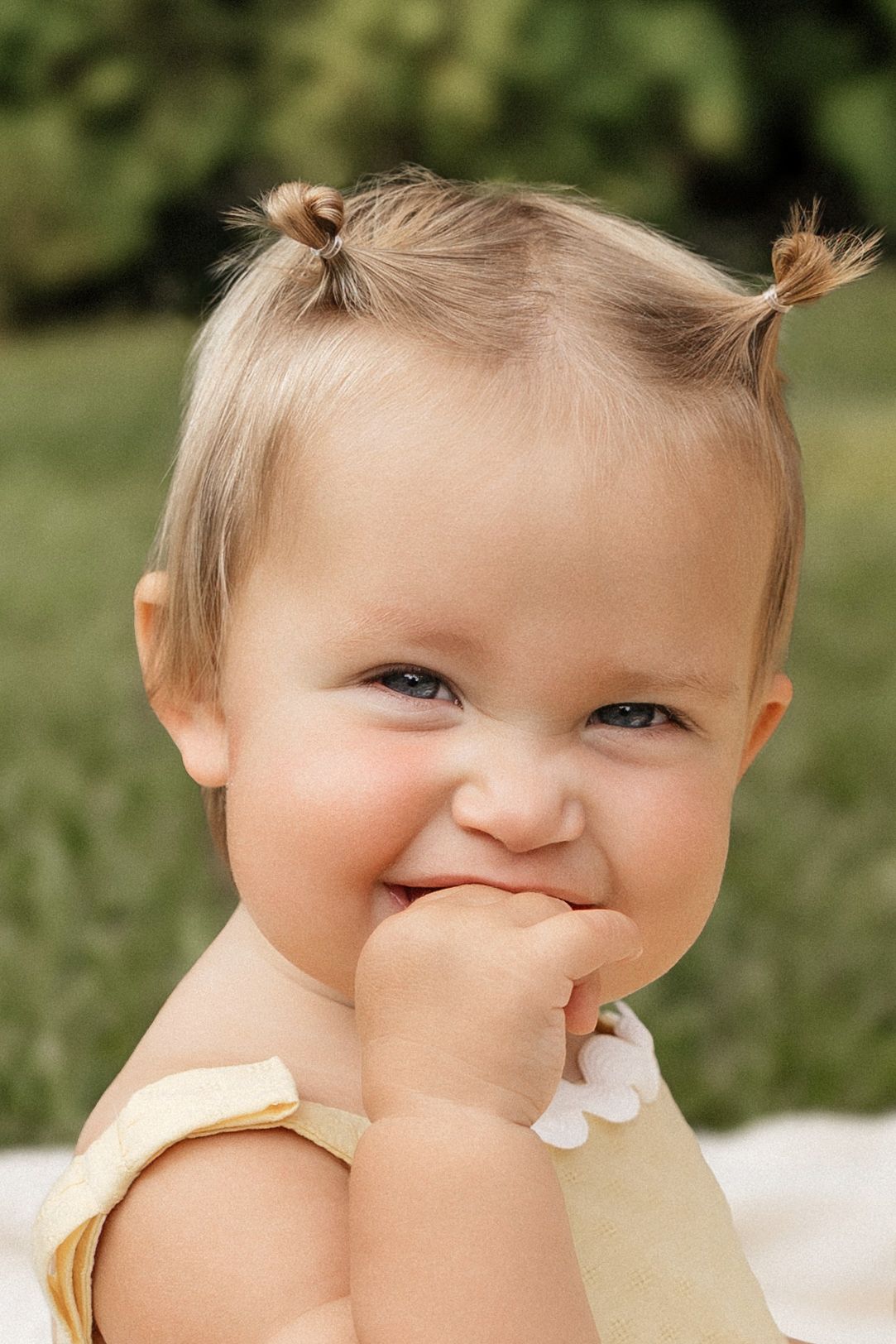 Adorable smiling baby girl with tiny pigtails wearing a yellow outfit, sitting outdoors on a sunny day.