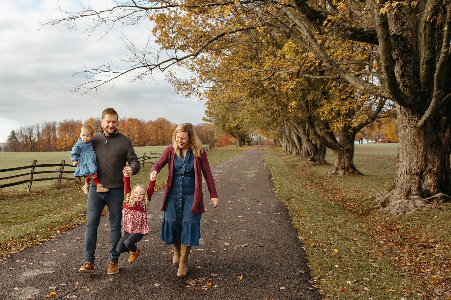 Family of four walking hand-in-hand down a tree-lined path during colorful autumn foliage season.