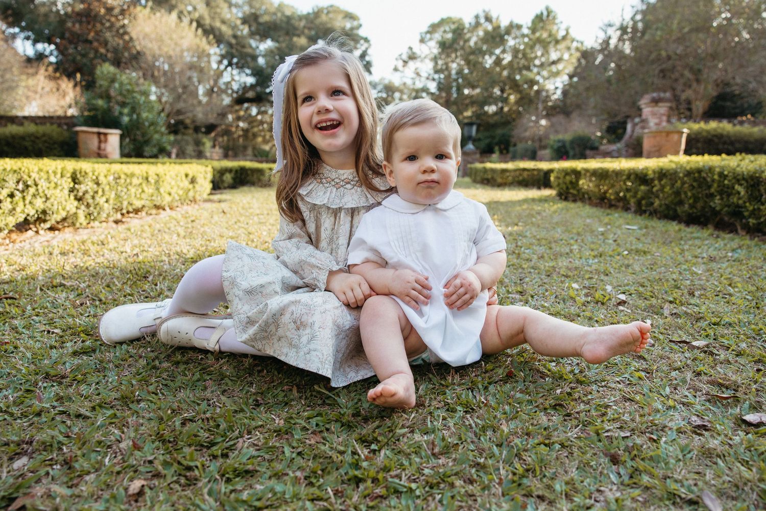 Two young siblings sitting on grass, older girl in floral dress posing with baby in white outfit.