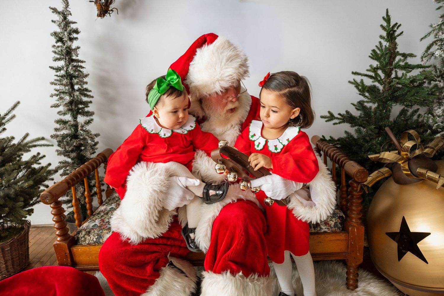 Two little girls in red dresses sit with Santa Claus examining gift in Christmas photo studio.