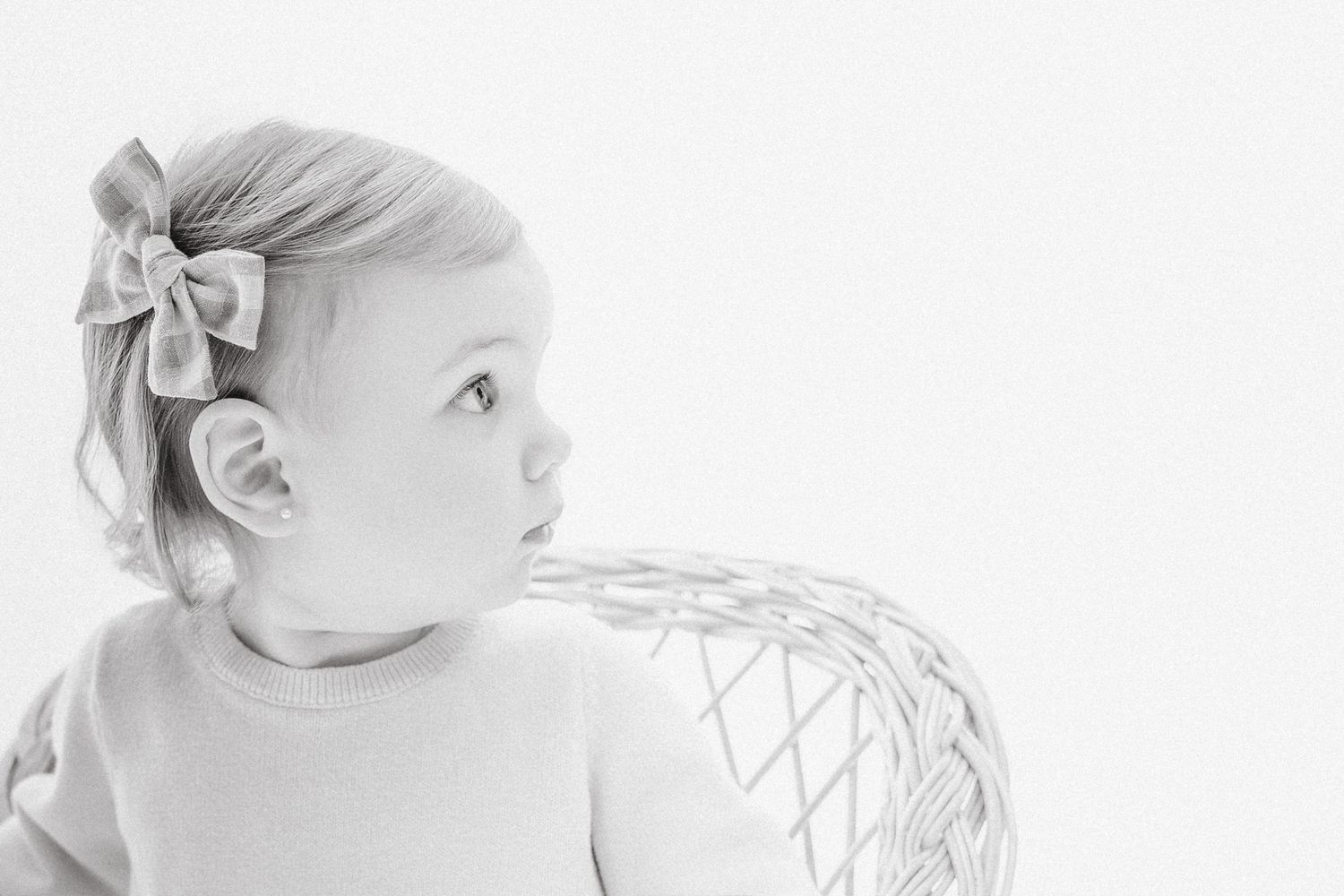 A black and white portrait of a baby girl with a bow headband sitting in a wicker chair.