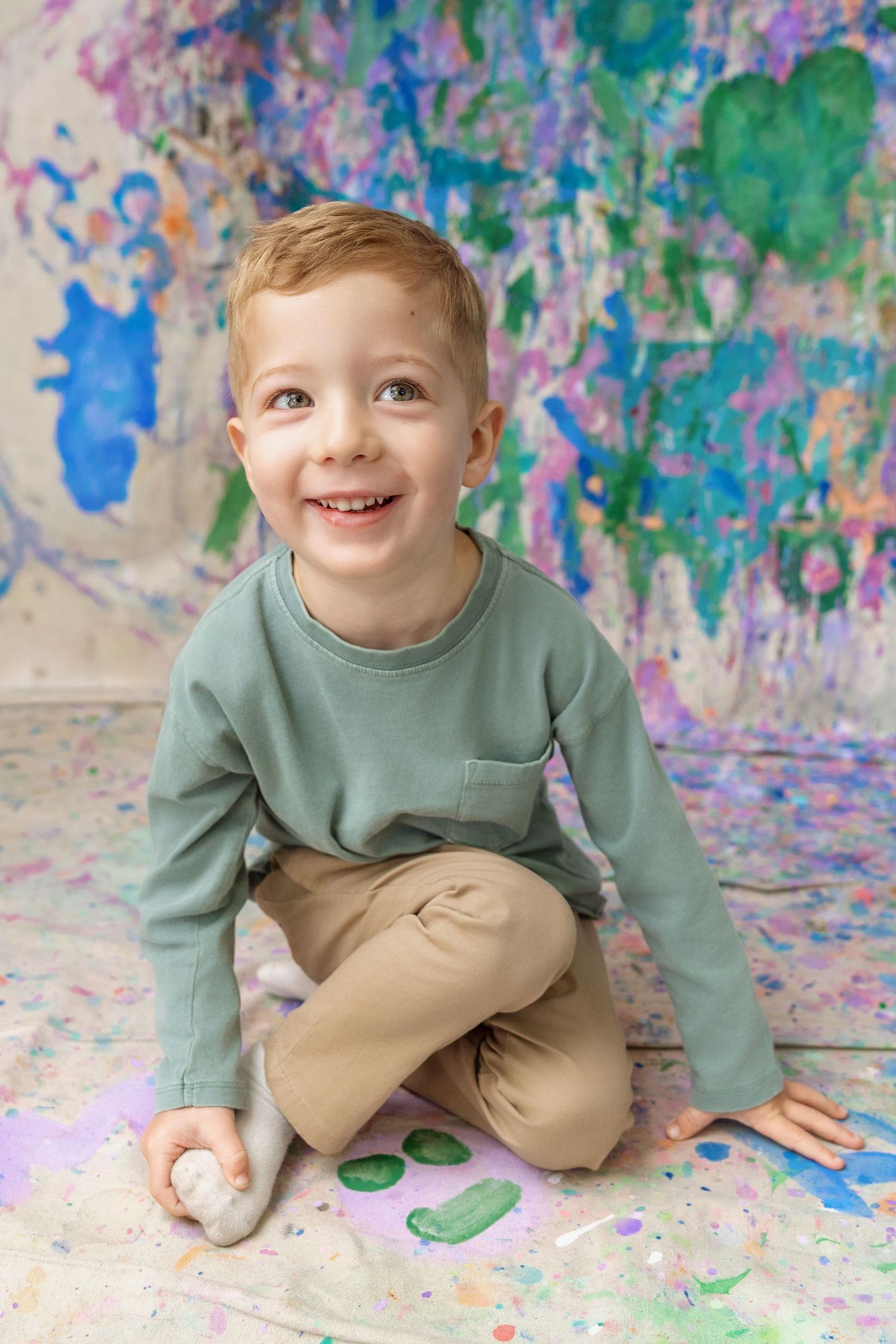 A smiling red-haired boy sits cross-legged on a paint-splattered floor before a colorful mural.