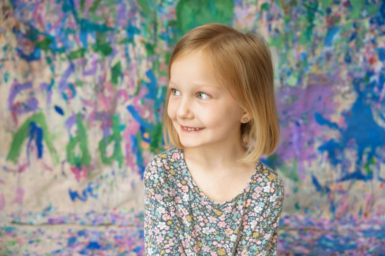 A smiling blonde girl in a floral dress poses before a vibrant multicolored painted wall.