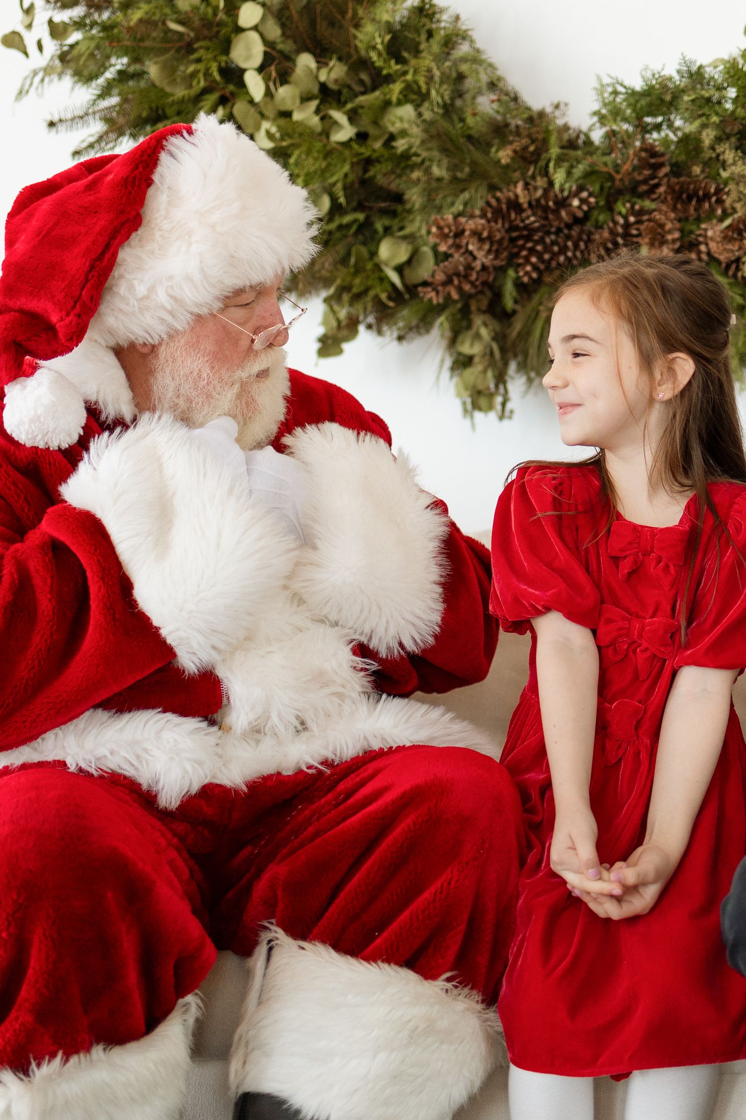 Santa Claus sits with a smiling young girl in a red velvet dress near a festive Christmas wreath.