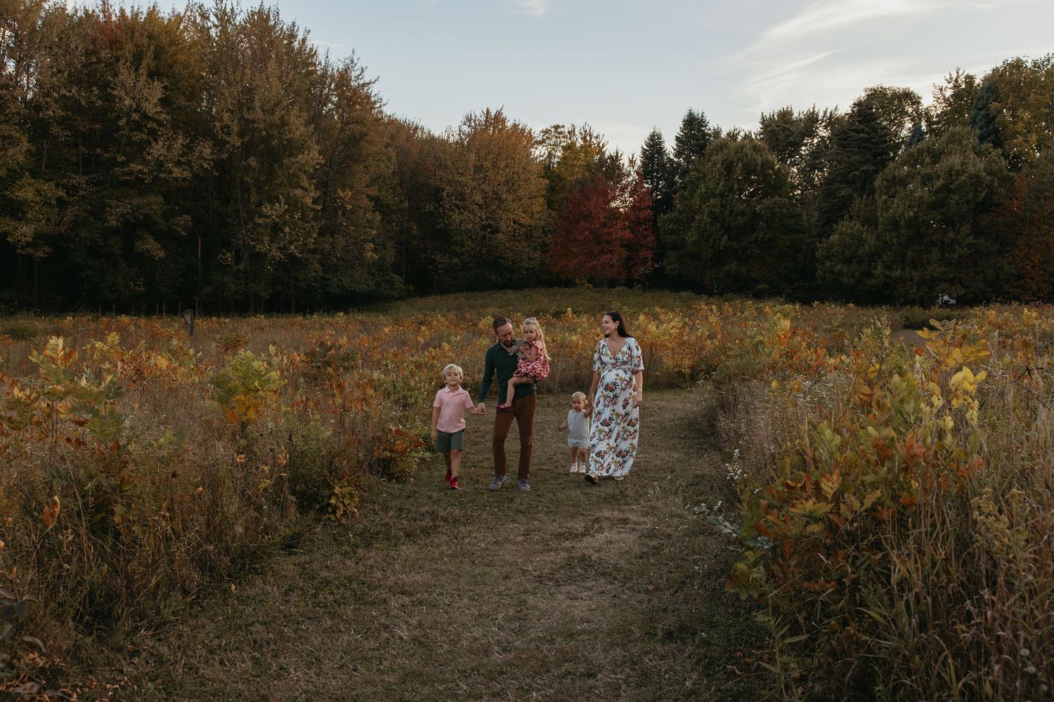 Family of five walking through an autumn meadow surrounded by colorful fall foliage trees.