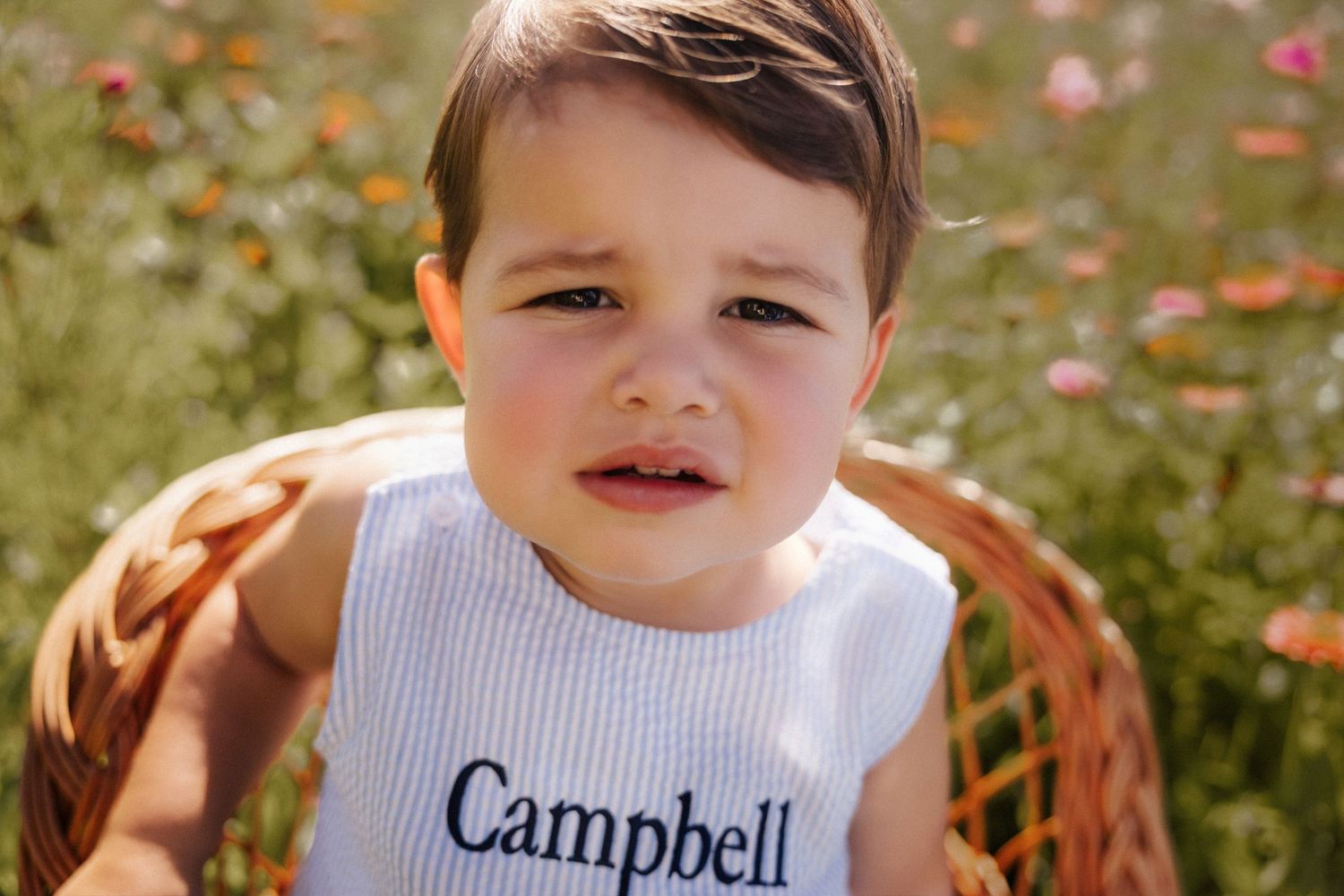 Toddler boy named Campbell sits in wicker chair wearing personalized white romper, surrounded by colorful wildflowers.