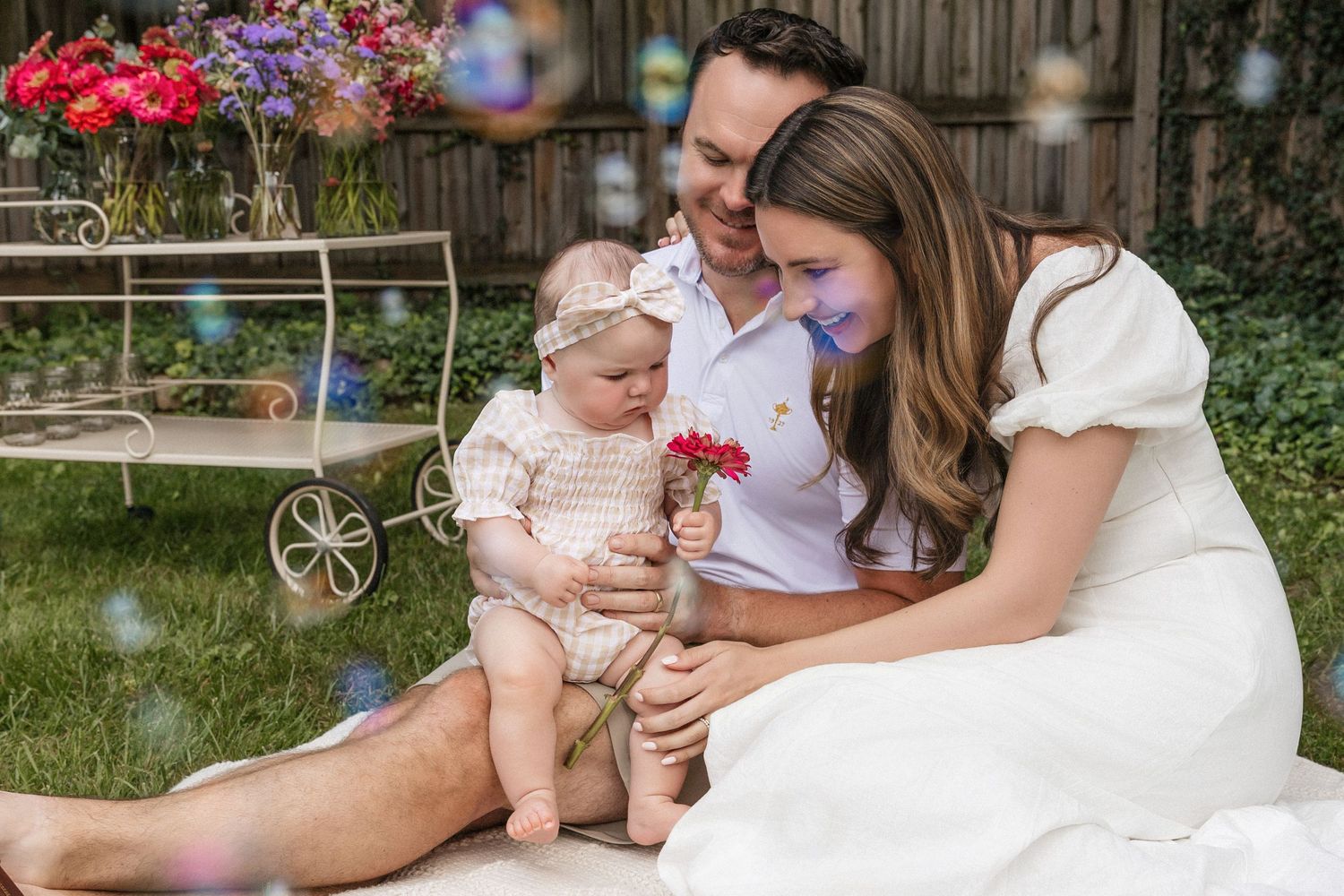 A happy family sits outdoors with bubbles, parents lovingly showing their baby girl a pink flower in a garden setting.