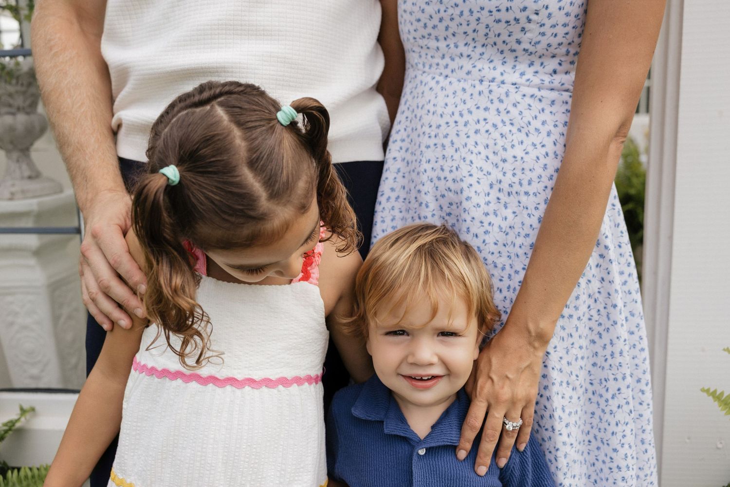 Two young children stand close to their parents outdoors, a girl in white dress and boy in blue shirt smiling.