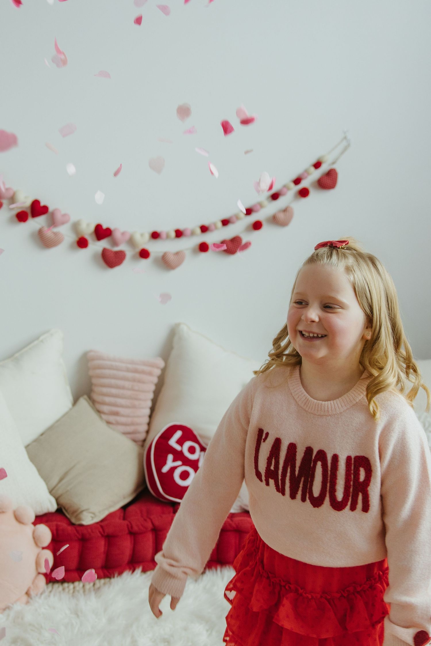 A cheerful blonde girl in a pink L'Amour sweater and red skirt laughs as heart confetti falls around her.