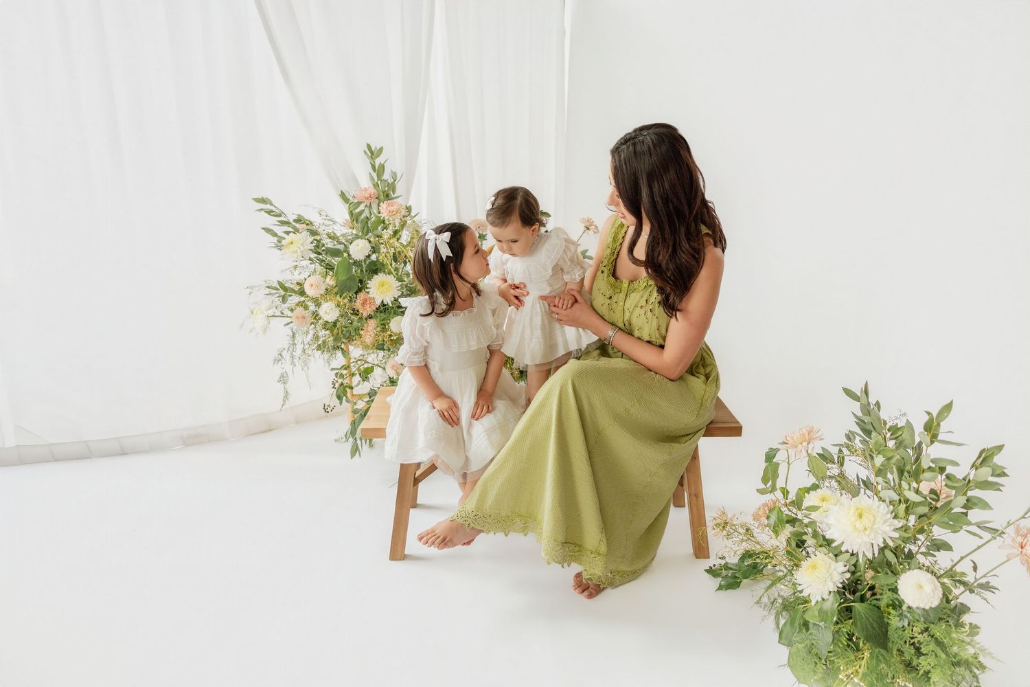 Mother and daughters in green and white dresses sitting on bench surrounded by flowers.