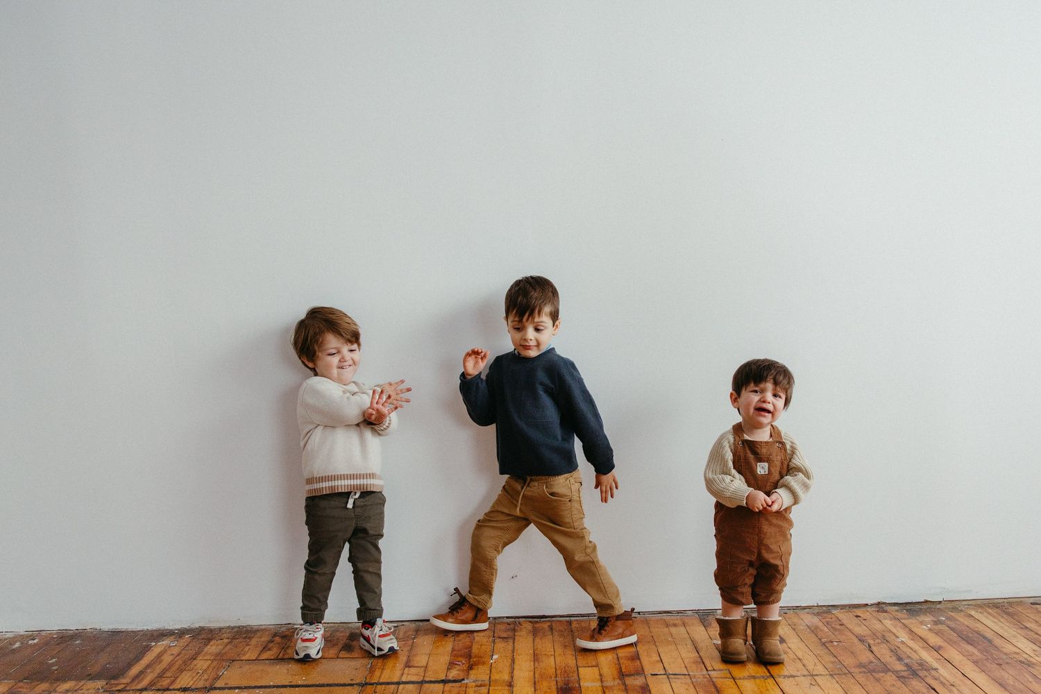 Three toddler boys playing and dancing against a white wall on a wooden floor.