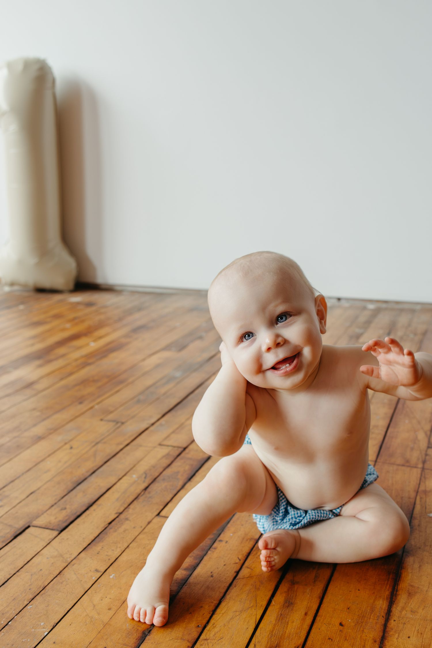 Happy baby in a blue diaper sitting on a hardwood floor smiling during an indoor lifestyle photography session.