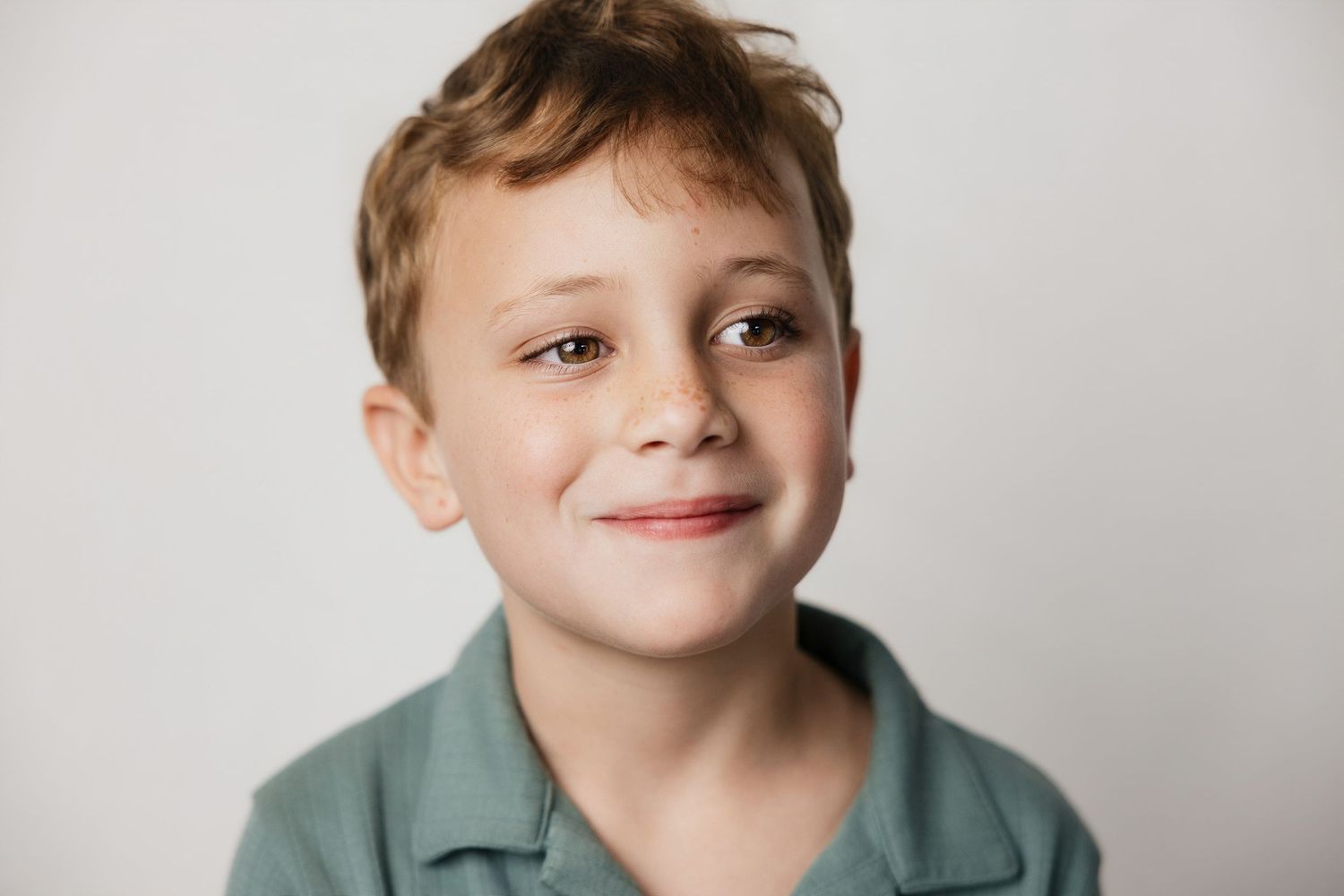 Smiling young boy with auburn hair wearing a green polo shirt against a white background.