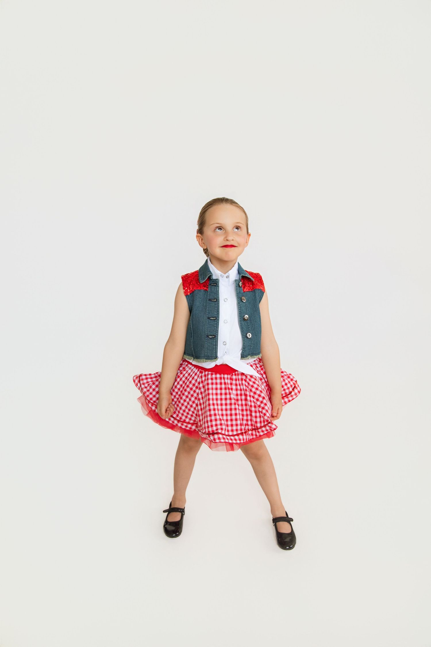 Young dance in red gingham skirt, denim vest, and black shoes poses confidently against white background.