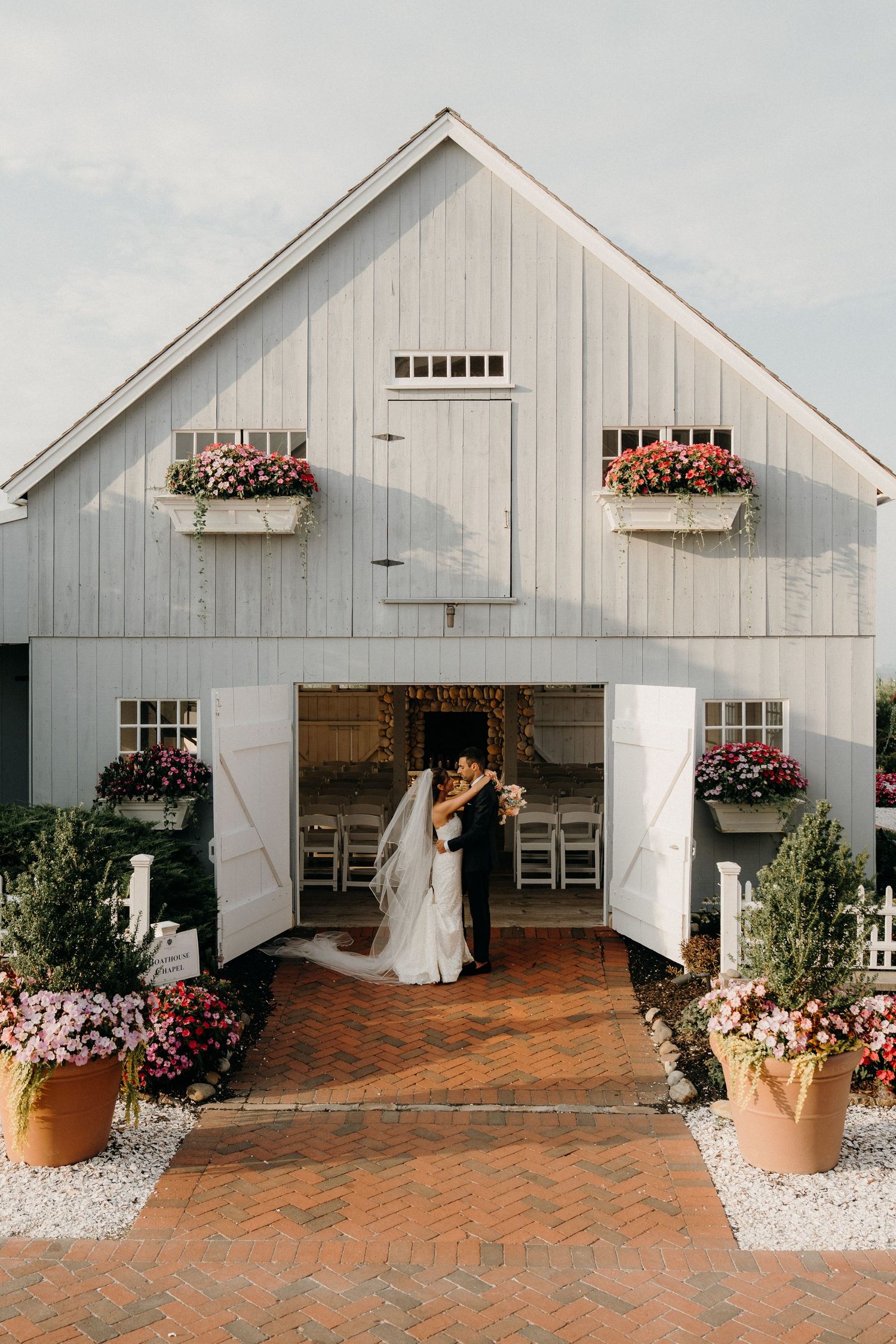 Bride and groom kissing at entrance of white barn wedding venue adorned with colorful flower boxes and potted plants.