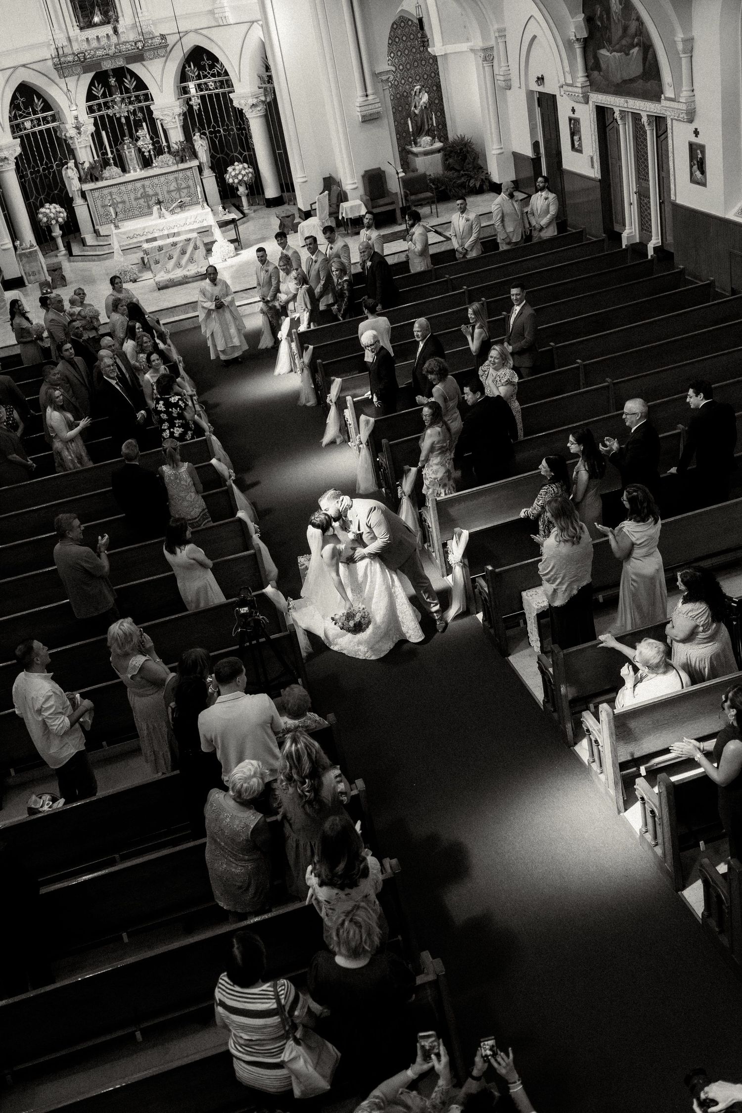 Black and white aerial view of a wedding couple kissing in church aisle, surrounded by standing guests in pews.